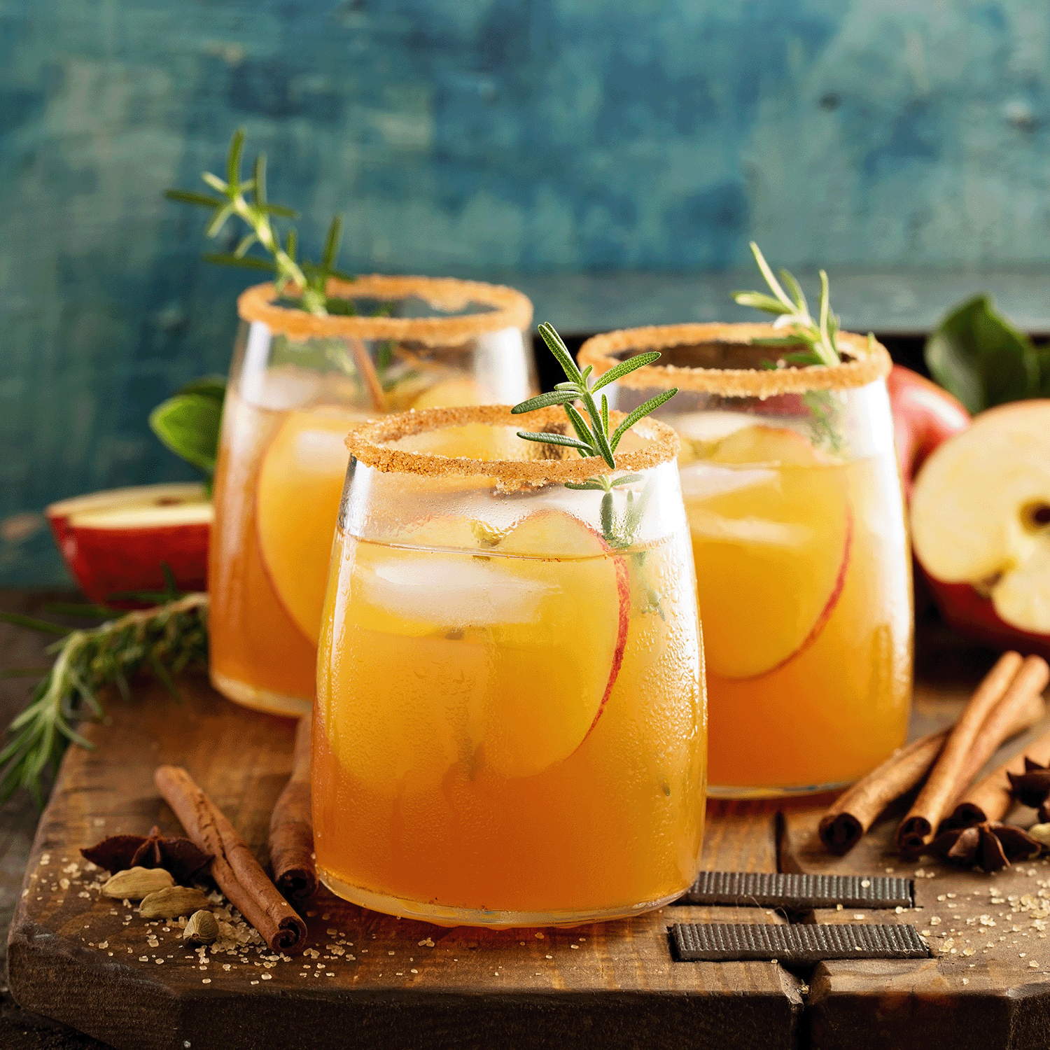 Three glasses of sparkling cider with apple slices, garnished with rosemary and cinnamon sticks, sit on a wooden board surrounded by spices and apples—evoking the cozy autumn atmosphere captured in the Tuscany Candle® SEASONAL Sparkling Cider: Bitters & Prosecco Scented Fall Wax Melt (2.5 oz).