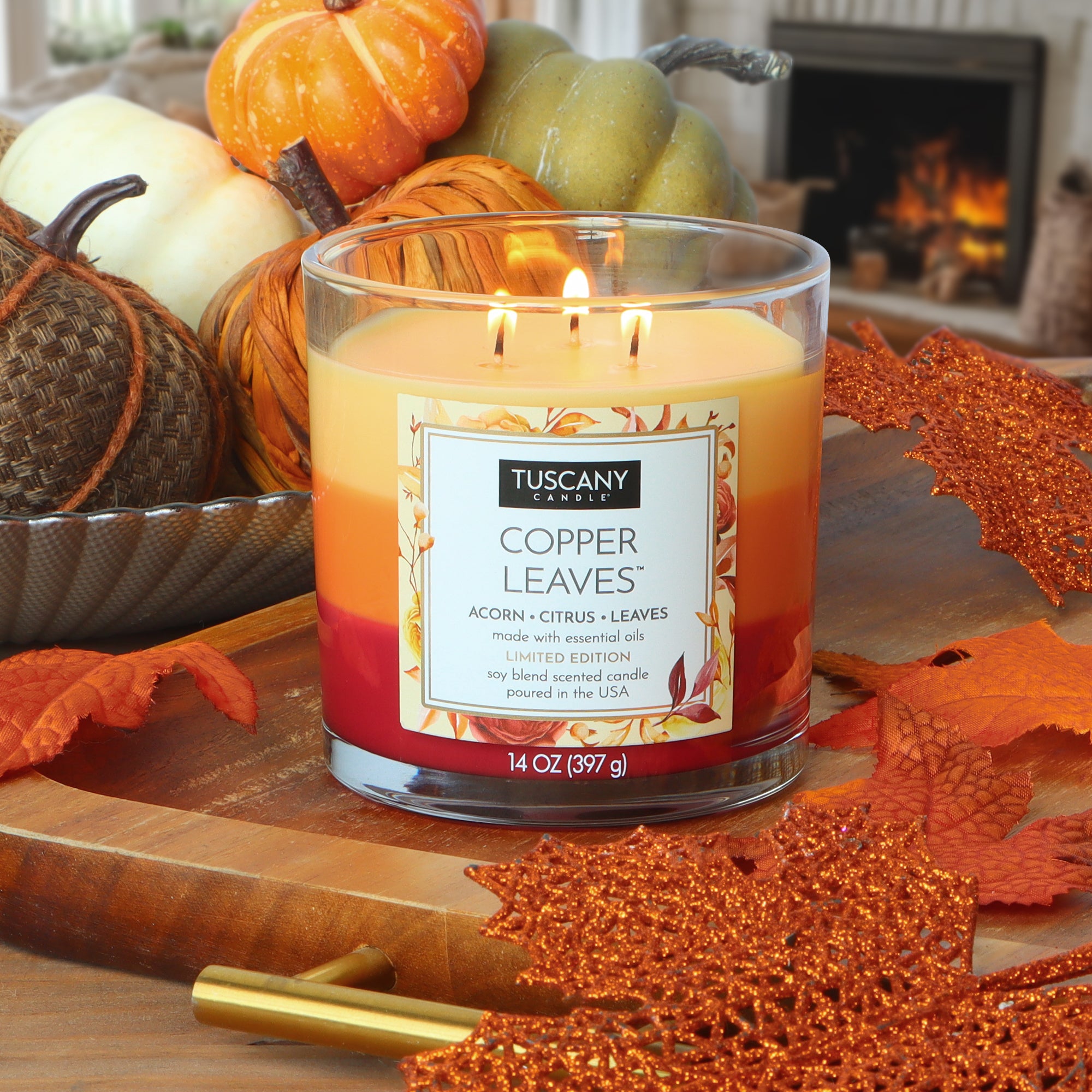 A lit Tuscany Candle® SEASONAL Copper Leaves: Acorns & Crushed Leaves Scented Fall Candle (14 oz) sits on a wooden table with pumpkins, acorns, and orange glittery leaves, a fireplace glowing softly in the background.