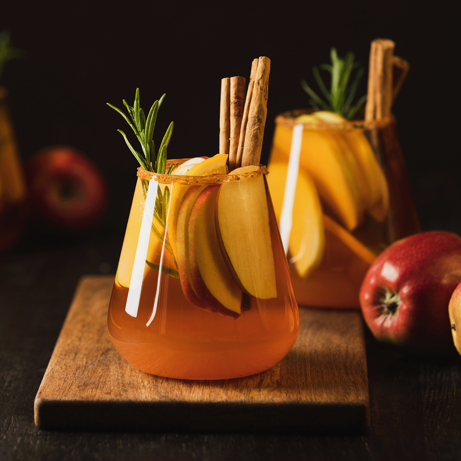 A glass of apple cider garnished with apple slices, rosemary, and cinnamon sticks, set on a wooden board alongside whole apples, captures the essence of the Tuscany Candle® SEASONAL Apple Cider: Clove Buds & Cinnamon Scented Fall Candle (14 oz)—a soy blend wax candle that creates a warm and cozy fall atmosphere.