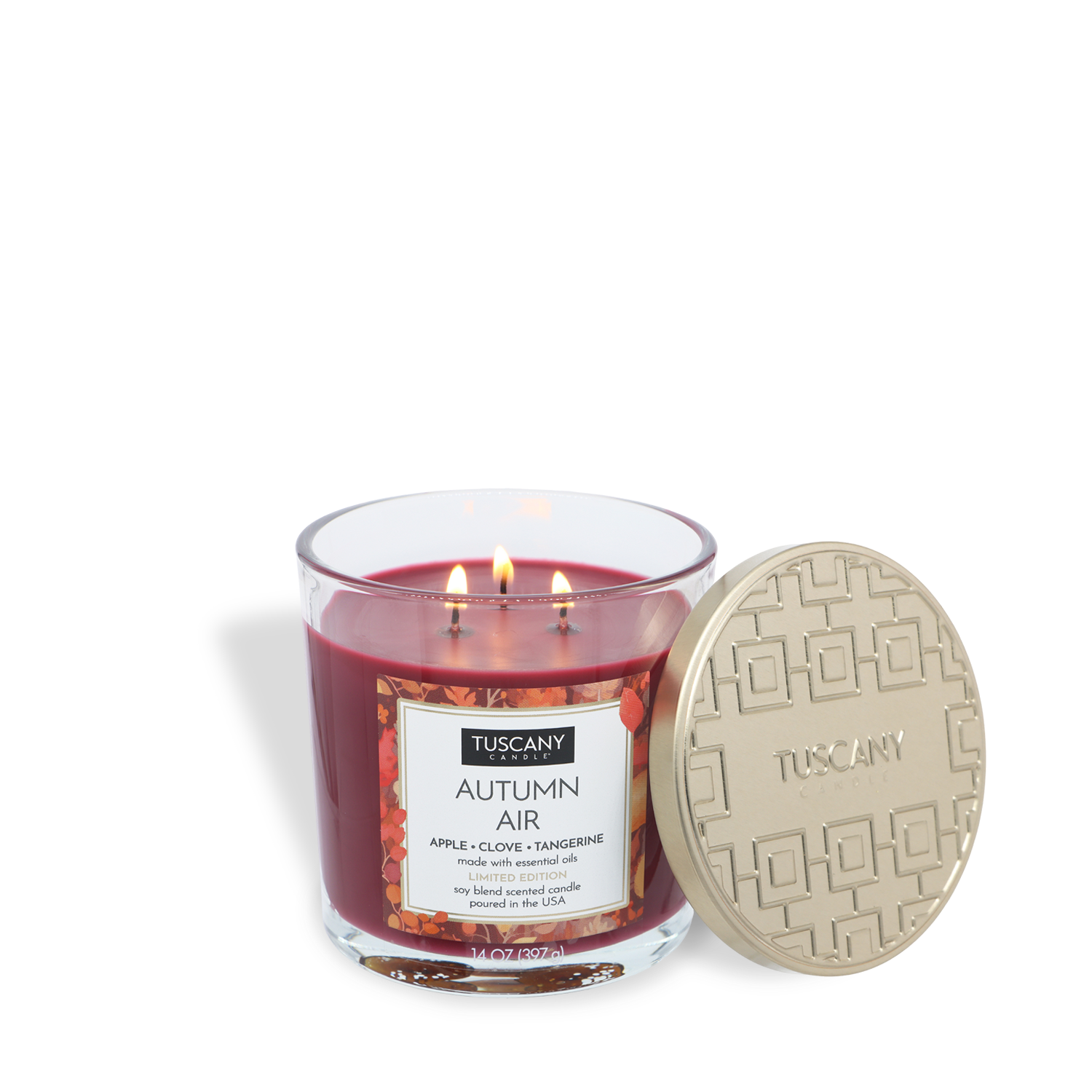 A red glass jar candle labeled “Autumn Air: Apple & Clove” by Tuscany Candle® SEASONAL, with three lit wicks and a gold-colored lid resting on its side. The candle weighs 14 oz and is perfect for fall.