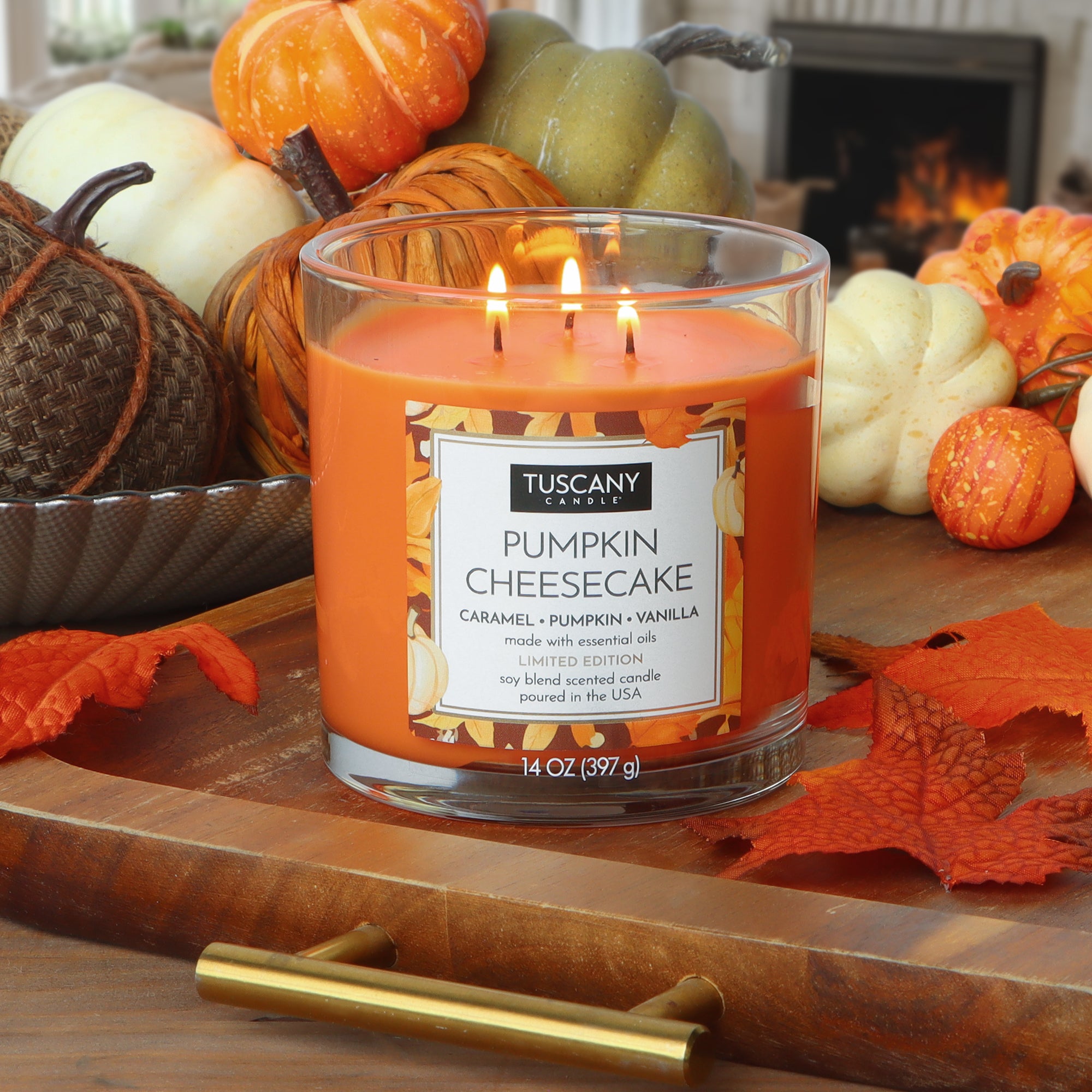 A lit 14 oz Tuscany Candle® SEASONAL sits on a wooden tray, featuring the Pumpkin Cheesecake: Caramel & Vanilla scent, surrounded by artificial pumpkins, leaves, and other fall decor in the background.