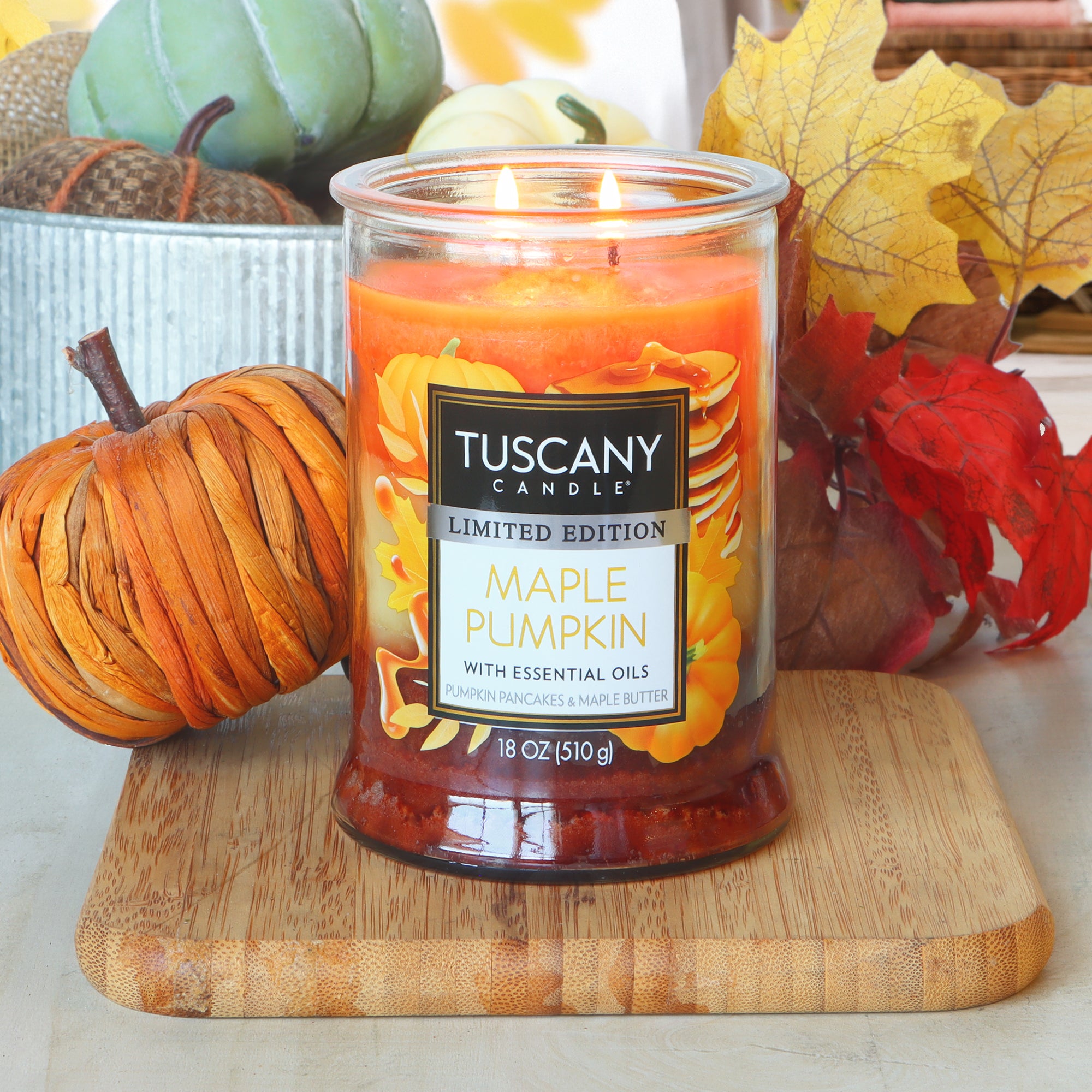 A lit Tuscany Candle® SEASONAL "Maple Pumpkin: Buttered Pancake Scented Fall Candle" (18 oz) rests on a wooden board, surrounded by artificial pumpkins, leaves, and a decorative tray.