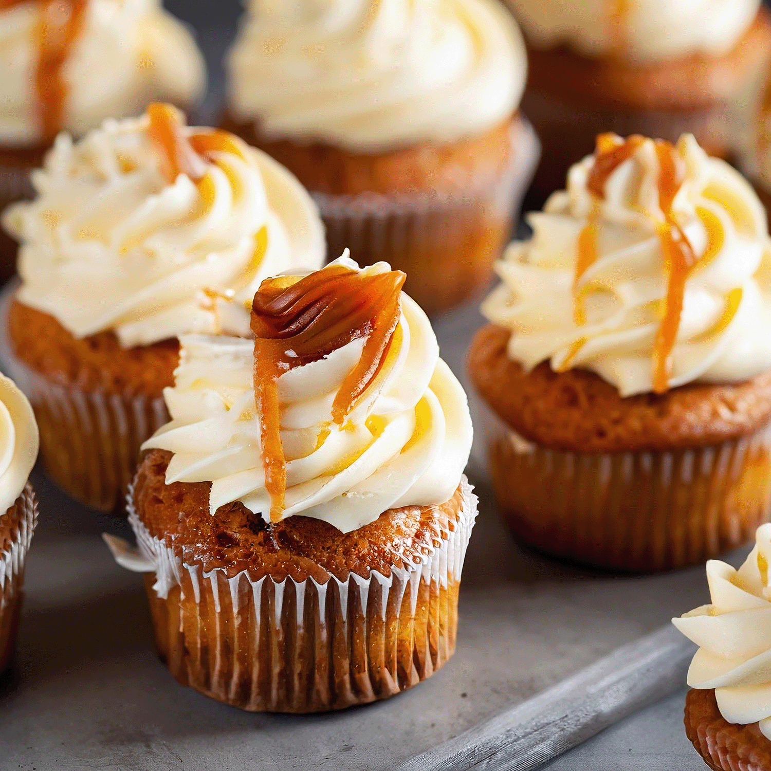 Cupcakes with swirled white frosting and caramel drizzle on top, arranged closely together on a gray surface—evocative of the warm, comforting aroma of Tuscany Candle® SEASONAL Maple Pumpkin: Buttered Pancake Scented Fall Candle (18 oz), perfect for capturing the essence of an autumn home fragrance.