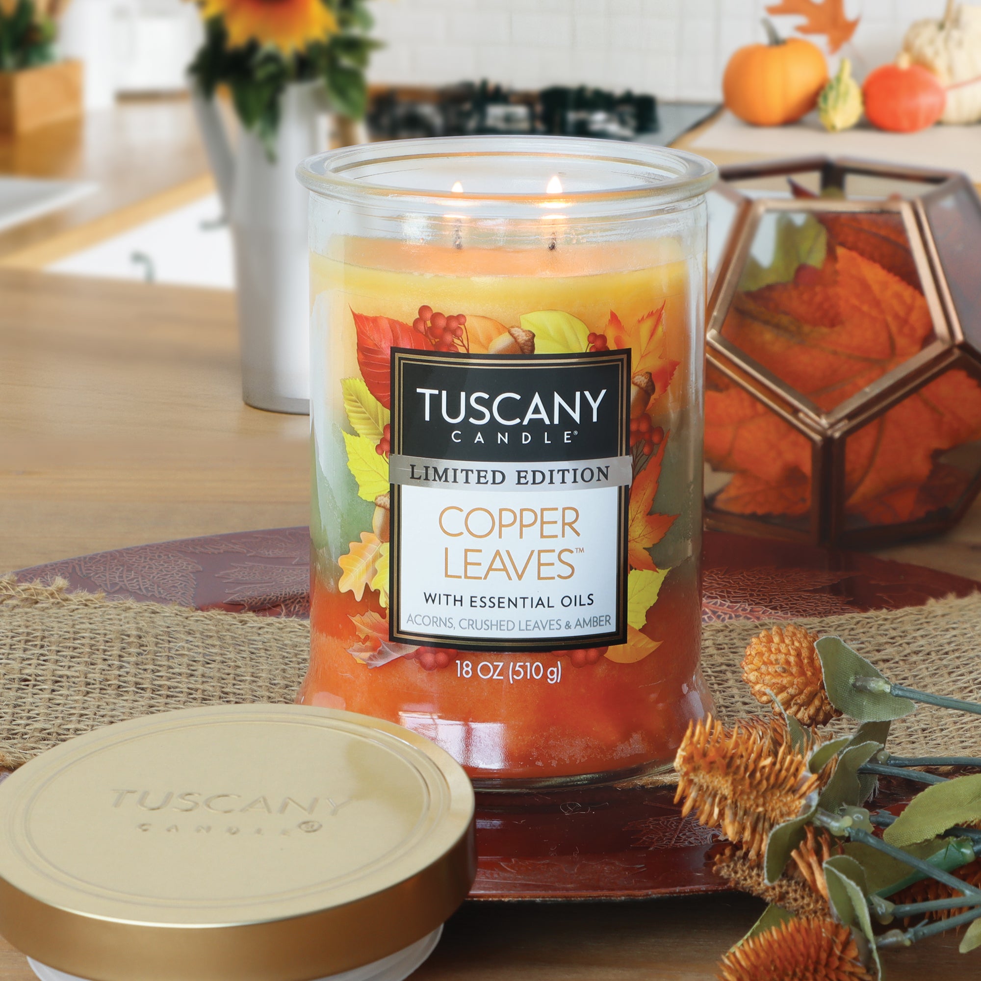 A lit Tuscany Candle® SEASONAL Copper Leaves®: Acorns & Crushed Leaves Scented Fall Candle (18 oz) sits on a kitchen counter, its premium marbled wax glowing warmly amidst autumn-themed decor and artificial orange leaves, creating the perfect autumn home fragrance.