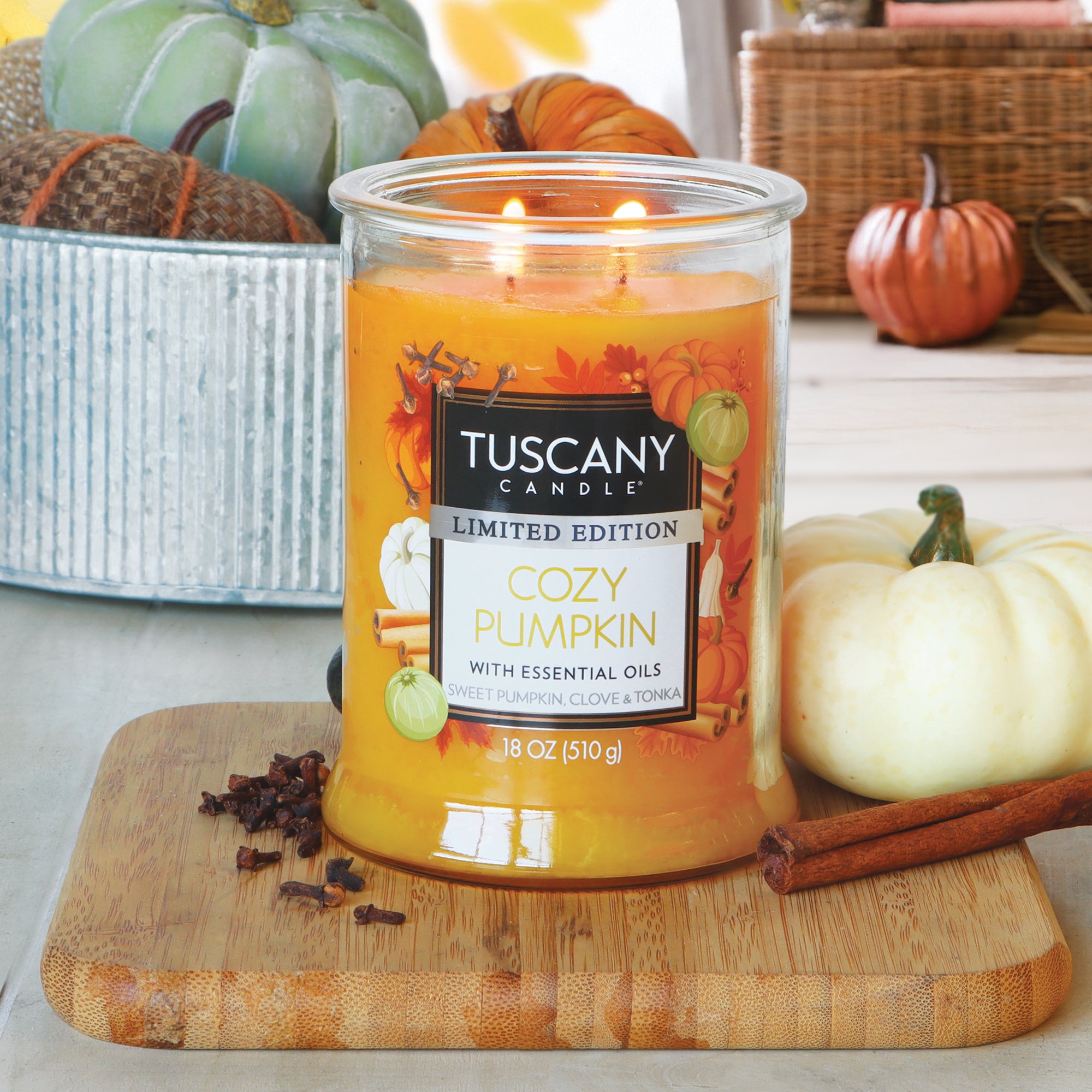 A lit Tuscany Candle® SEASONAL "Cozy Pumpkin: Sweet Pumpkin & Clove" 18 oz candle sits on a wooden board with cinnamon sticks, cloves, and a white pumpkin, surrounded by autumn decor.