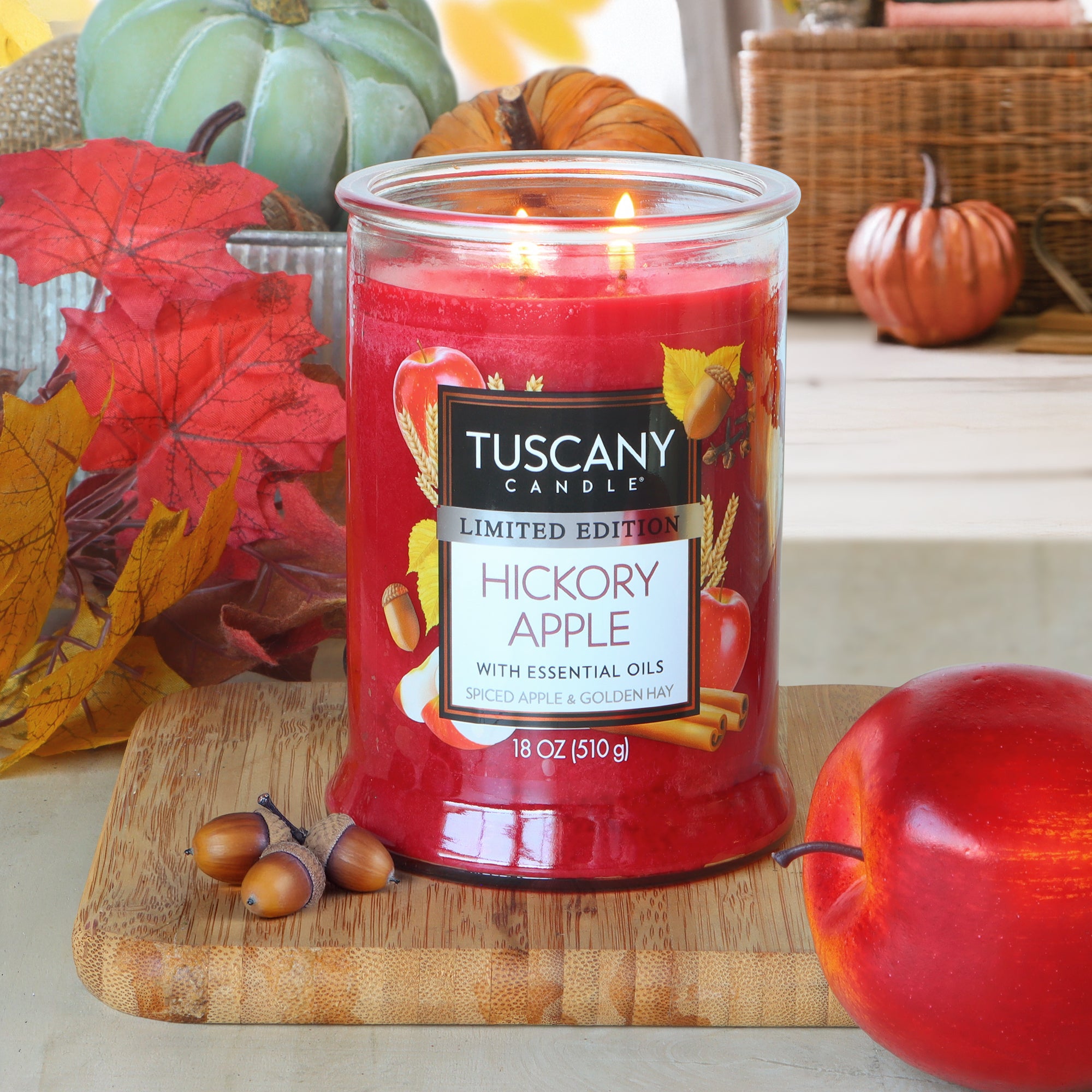 A lit Tuscany Candle® SEASONAL "Hickory Apple: Spiced Apple & Golden Hay" (18 oz) rests on a wooden board with acorns, an apple, and autumn decor like leaves, pumpkins, and a basket.
