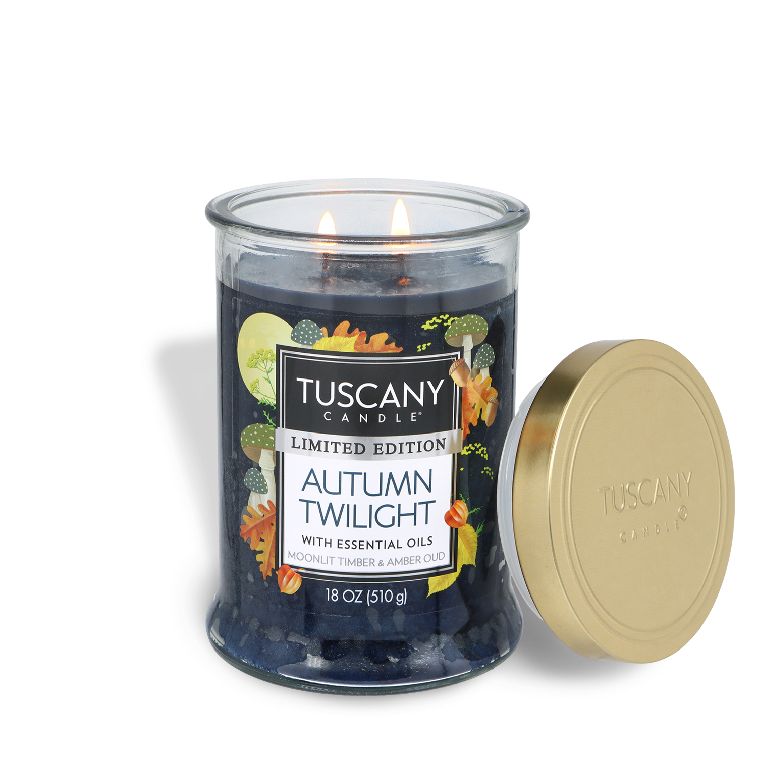 An 18 oz glass jar Tuscany Candle® SEASONAL “Autumn Twilight: Moonlit Timber & Amber Oud” candle with two lit wicks, adorned with autumn leaves and a gold lid on the right.