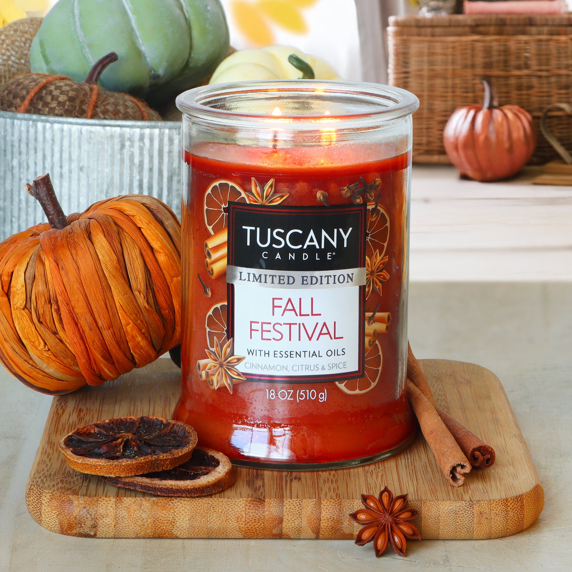 A lit Tuscany Candle® SEASONAL "Fall Festival: Cinnamon & Citrus Scented Fall Candle" (18 oz) sits on a wooden board with dried citrus, cinnamon stick, and star anise among pumpkin-themed fall decorations.