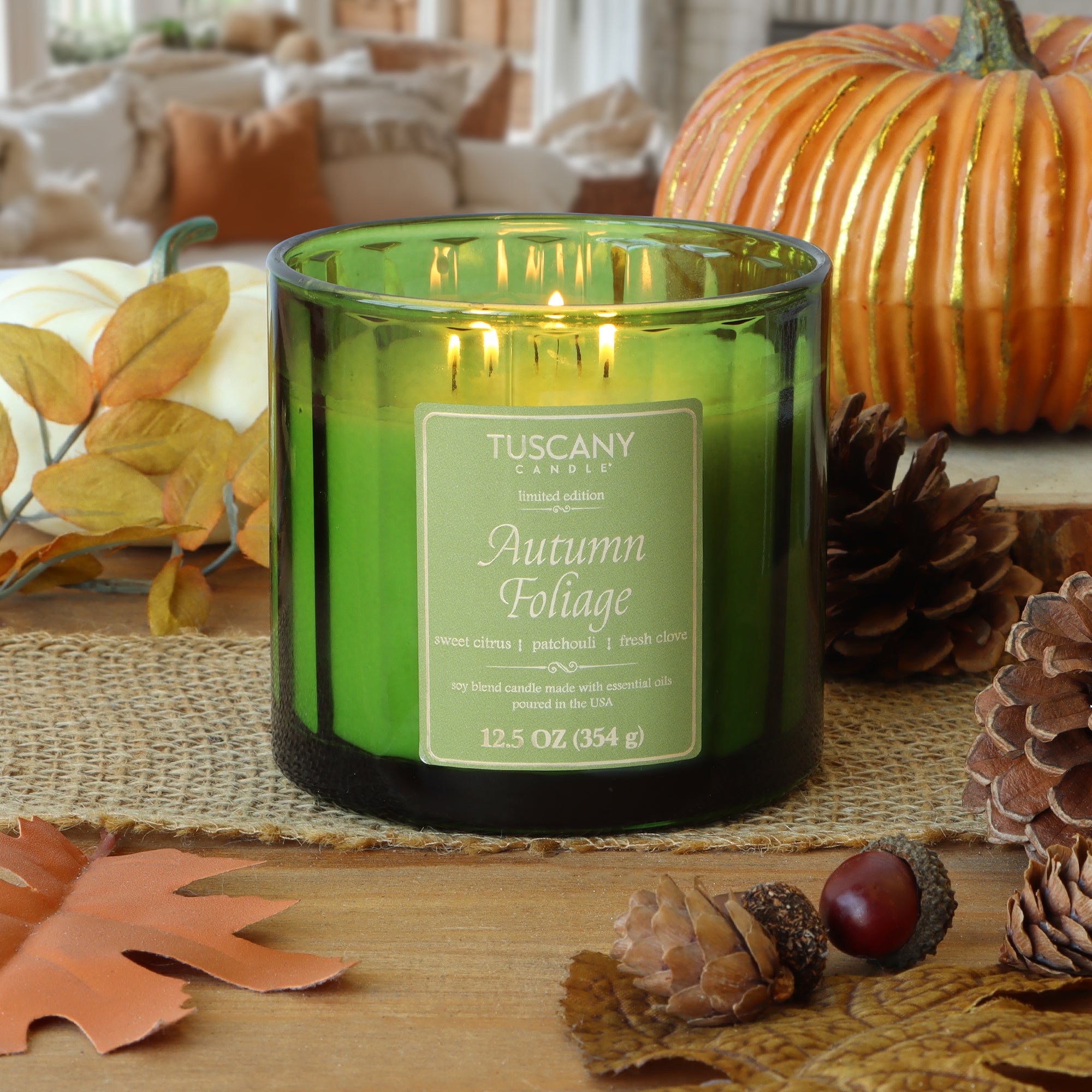A lit Tuscany Candle® SEASONAL Autumn Foliage: Citrus & Patchouli Scented Fall Candle (12.5 oz) sits on a wooden table, surrounded by autumn leaves, pine cones, and a pumpkin, filling the space with cozy fall fragrance.