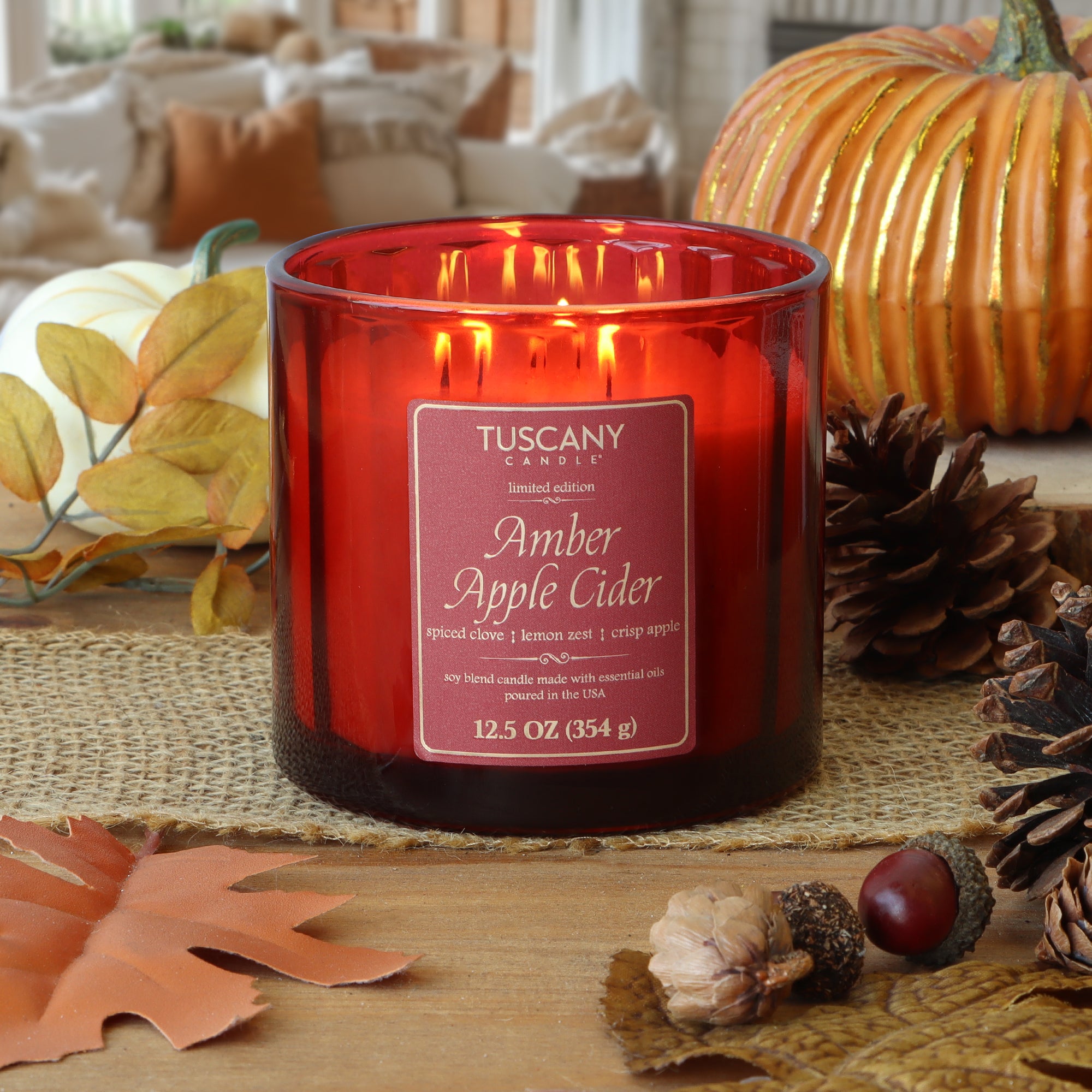 A Tuscany Candle® SEASONAL "Amber Apple Cider: Clove & Lemon Zest" 12.5 oz red glass fall candle sits among pinecones, pumpkins, and autumn decor, filling the cozy living room with a warm seasonal fragrance.