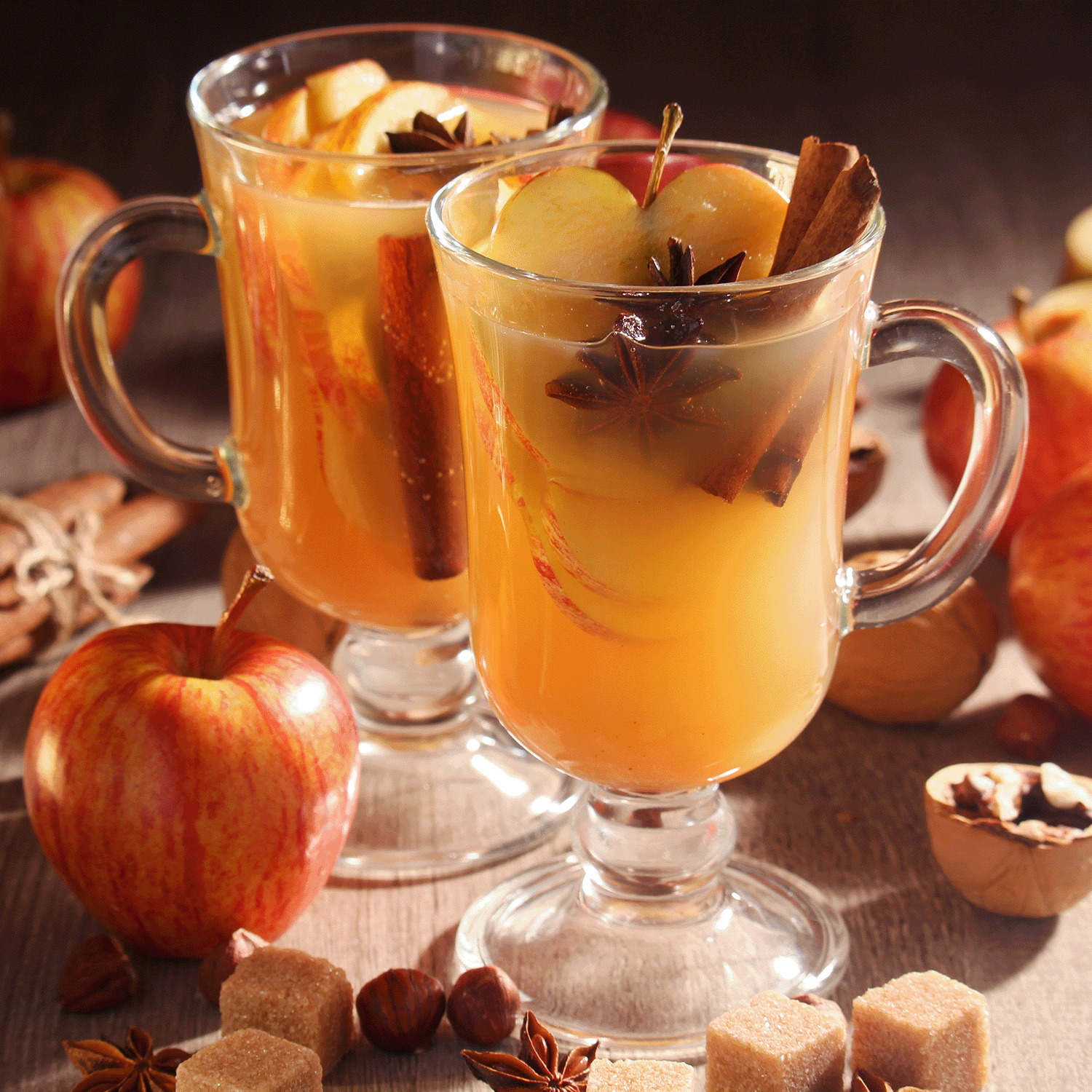Two glass mugs of spiced apple cider, topped with apple slices, cinnamon sticks, and star anise, rest on a wooden surface amidst apples, walnuts, spices, and sugar cubes—evoking the warm and inviting ambiance created by Tuscany Candle® SEASONAL’s Amber Apple Cider: Clove & Lemon Zest Scented Fall Candle (12.5 oz).