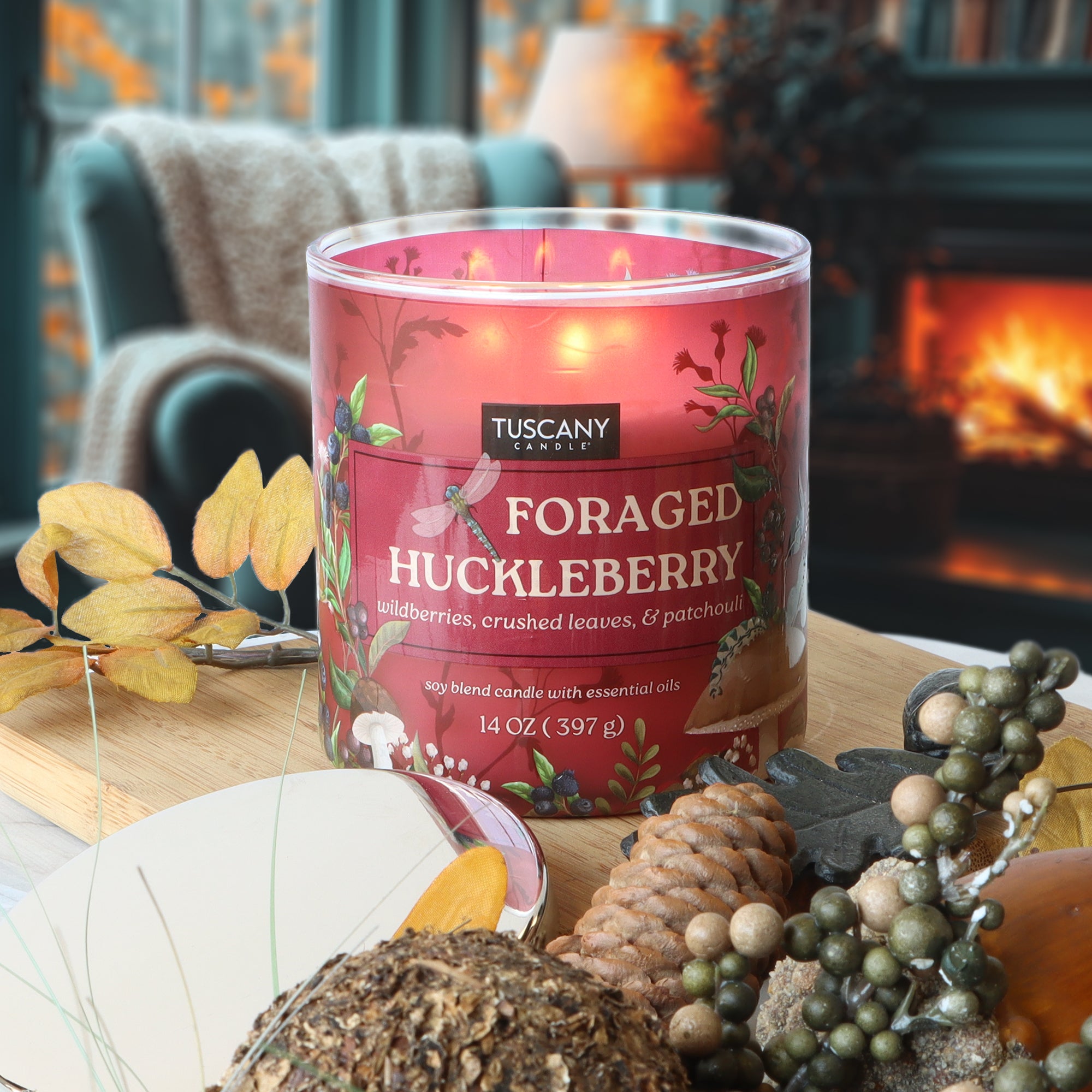 A lit Tuscany Candle® SEASONAL "Fireside Black Cherry: Vanilla & Praline" scented fall candle (14 oz) sits on a table with pinecones, berries, and dried leaves, fireplace and chair in the background.