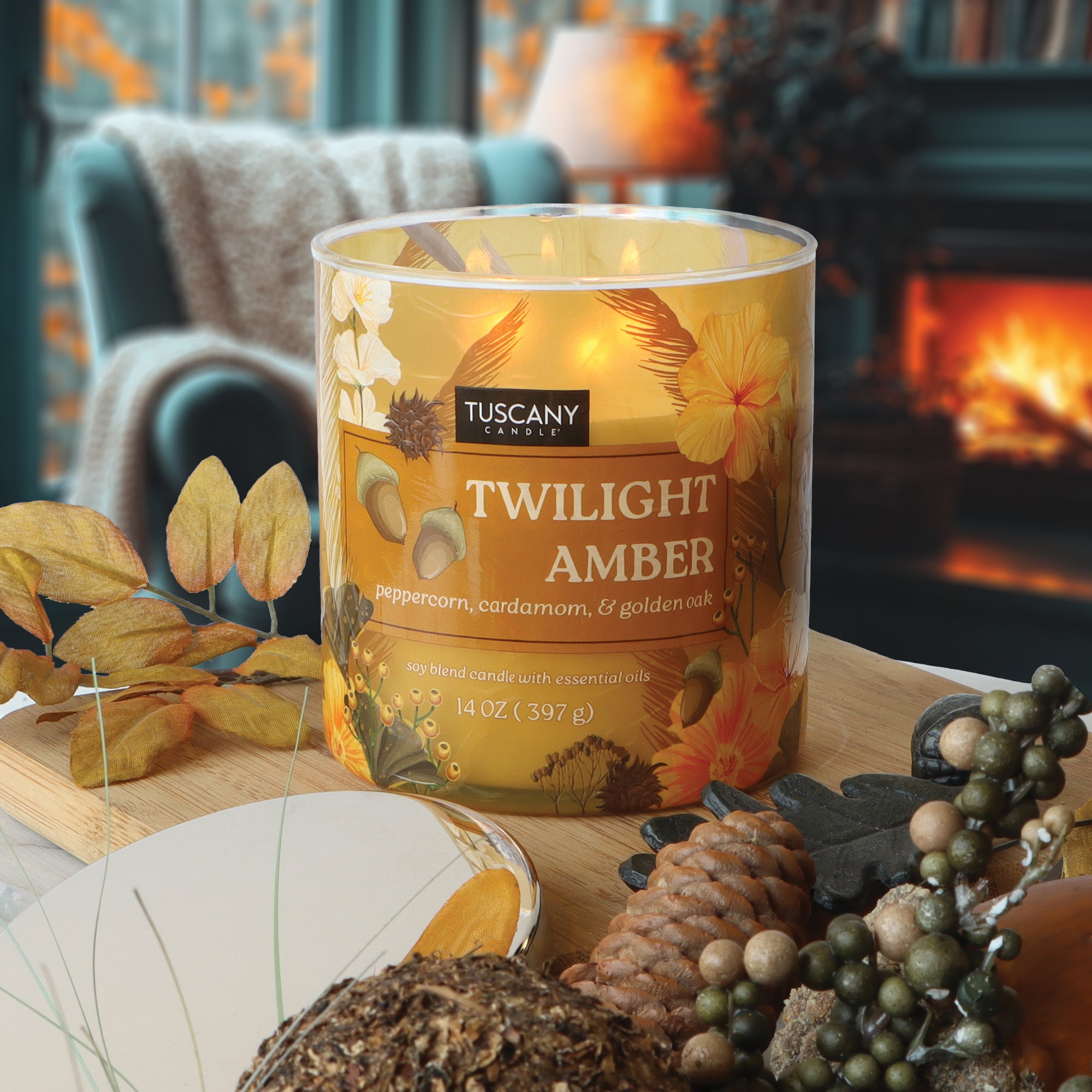 A lit Tuscany Candle® SEASONAL "Twilight Amber: Peppercorn & Cardamom Scented Fall Candle" (14 oz) sits on a table with autumn decor in front of a fireplace.