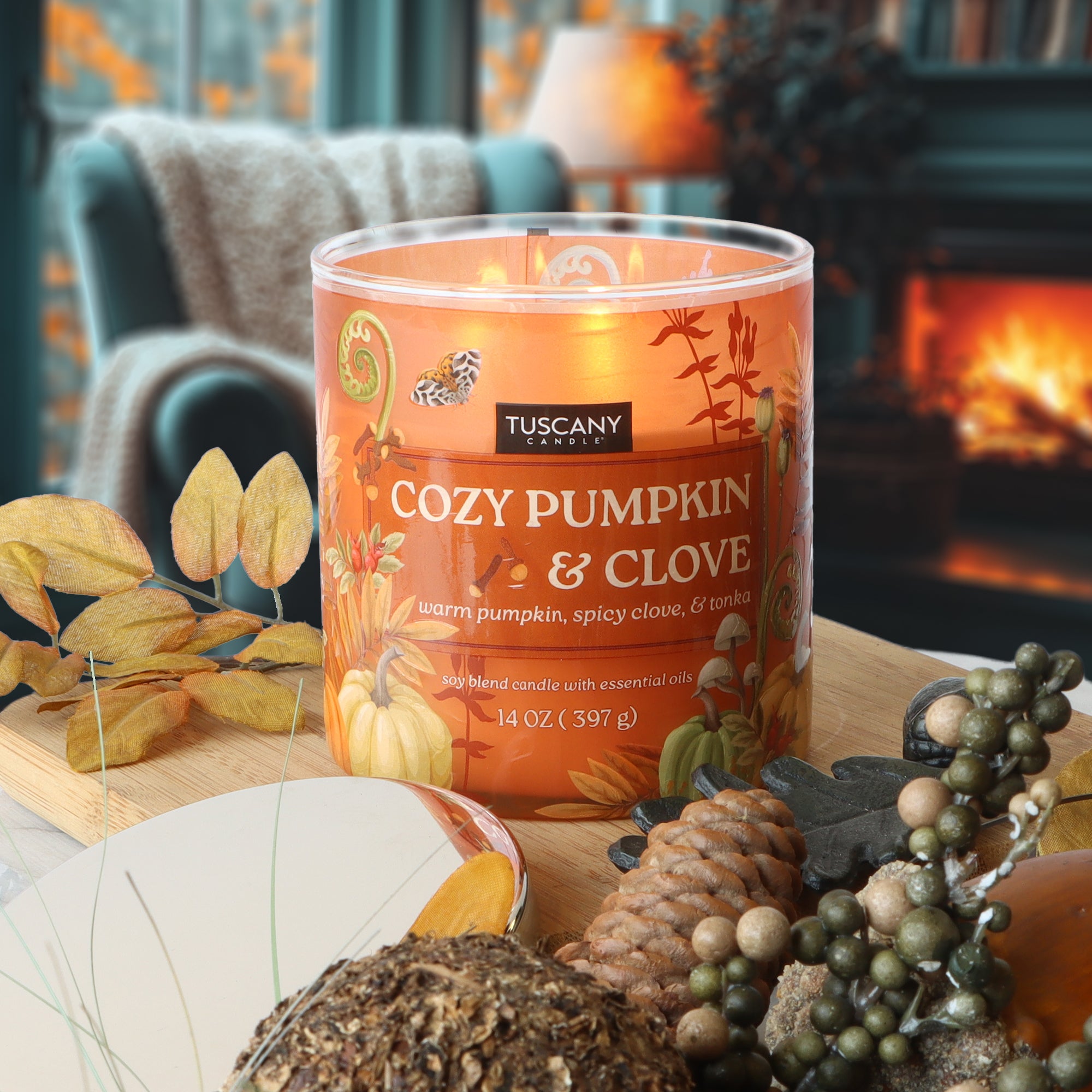 A Tuscany Candle® SEASONAL "Cozy Pumpkin & Clove: Warm Tonka Scented Fall Candle" (14 oz) sits on a table with autumn decor, a fireplace, and an armchair in the background.