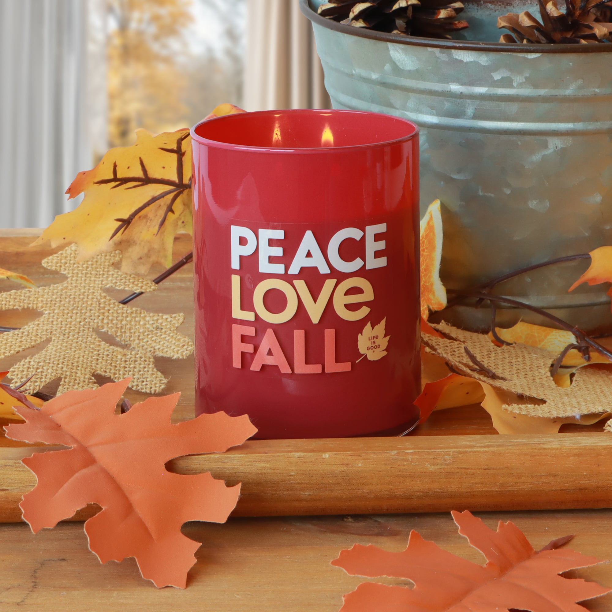 A Tuscany Candle® SEASONAL "Peace, Love, & Fall: Cinnamon & Spice" 13 oz candle by Life Is Good® sits on a wooden surface with artificial autumn leaves and a metal pot of pinecones.