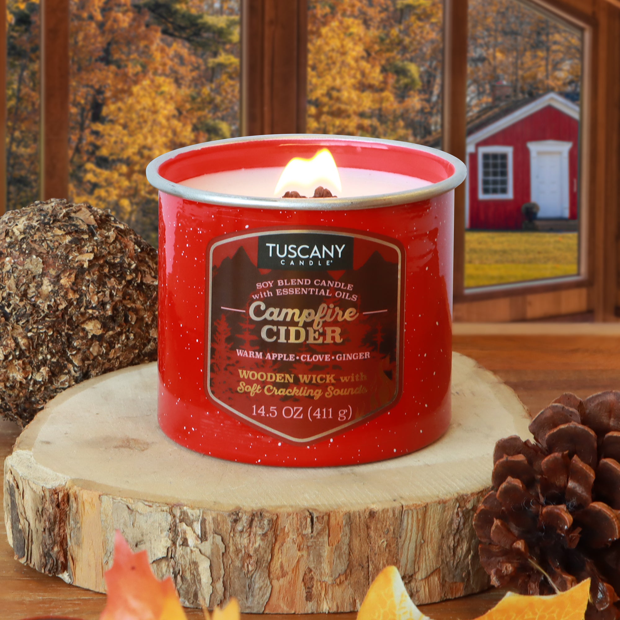 A Tuscany Candle® SEASONAL "Campfire Cider: Warm Apple & Clove" fall wooden wick candle (14.5 oz) sits on a wooden surface with pine cones and autumn decor, while a red barn is visible outside the window.