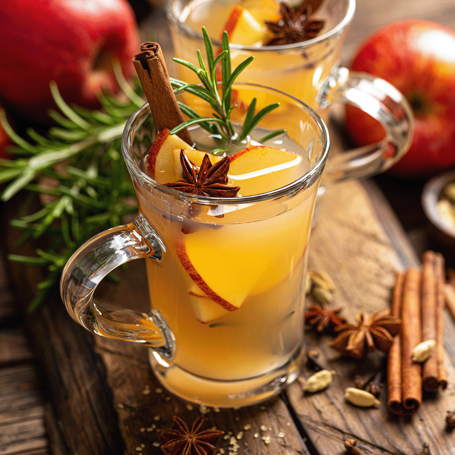 Two glass mugs of spiced apple cider garnished with apple slices, star anise, rosemary, and cinnamon sticks rest on a wooden surface surrounded by apples and spices—a cozy scene that captures the warm glow and inviting aroma of Tuscany Candle® SEASONAL’s Campfire Cider: Warm Apple & Clove Scented Fall Wooden Wick Candle (14.5 oz).