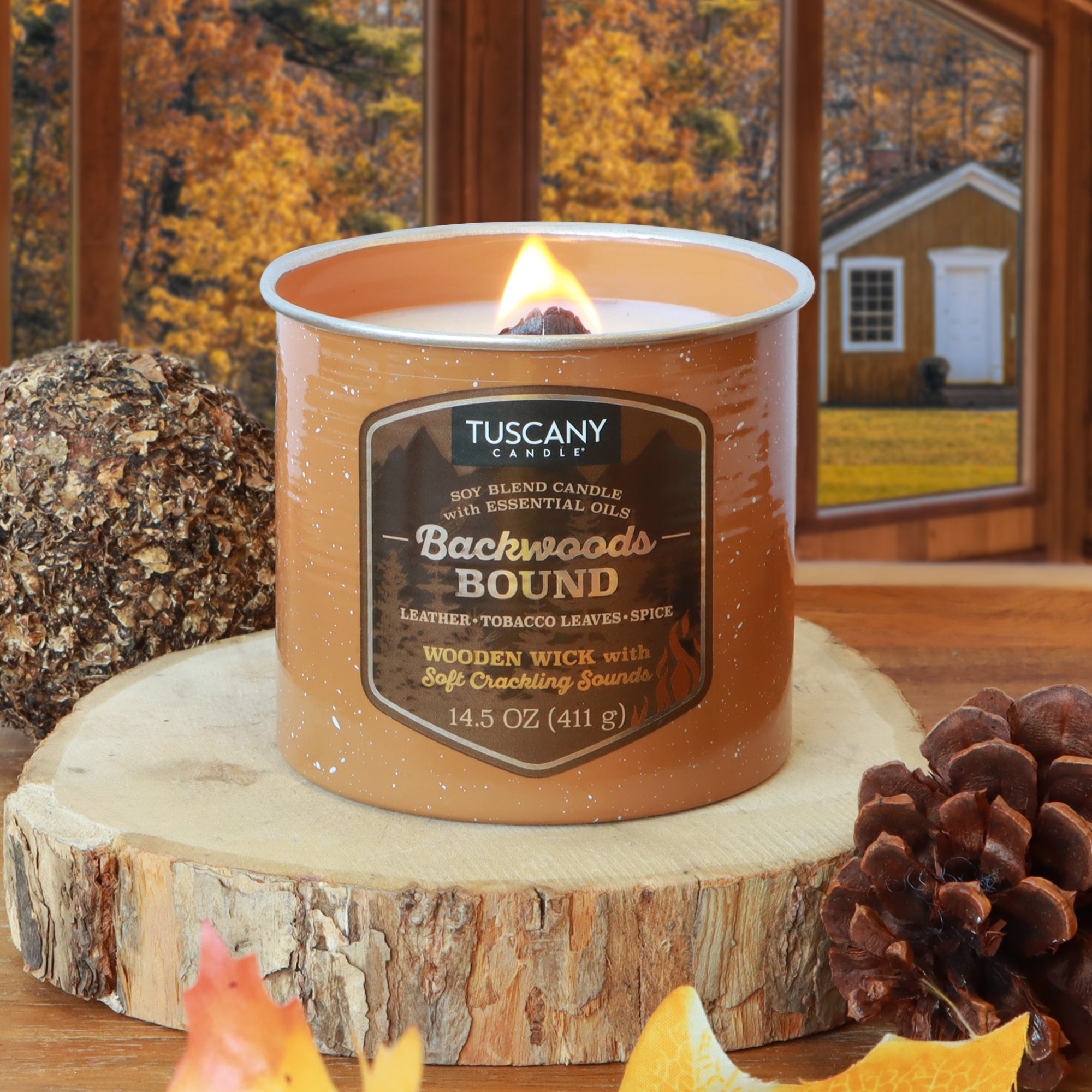 A lit Tuscany Candle® SEASONAL “Backwoods Bound: Leather & Tobacco Scented Fall Wooden Wick Candle (14.5 oz)” rests on a tree stump with pinecones, leaves, and a cabin visible—evoking cozy fall nights by the fire.