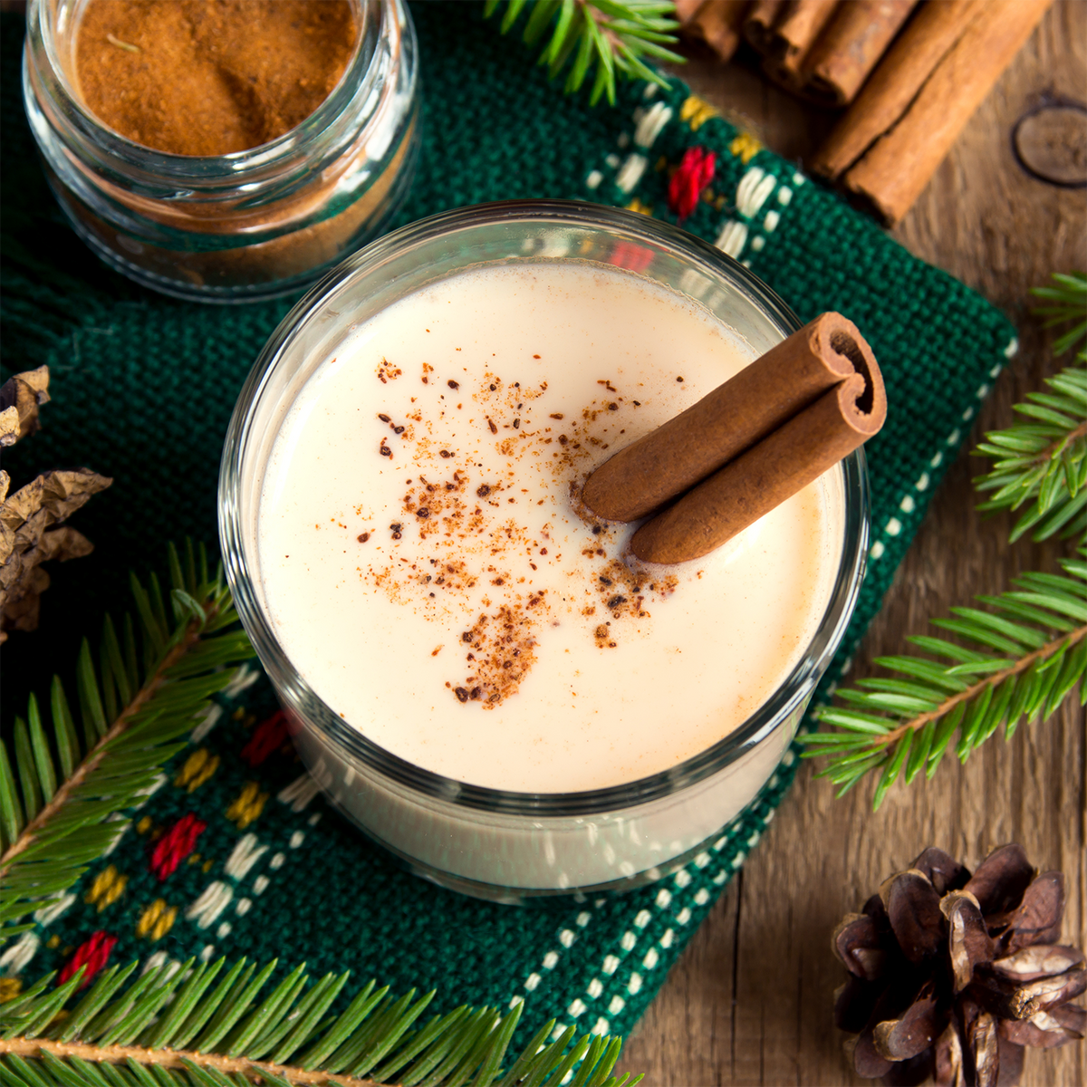 A glass of creamy Yuletide Vanilla: Smooth Rum & Clove Scented Holiday Wax Melt (2.5 oz) garnished with cinnamon sticks, set on a green knitted mat with pine needles and cinnamon.