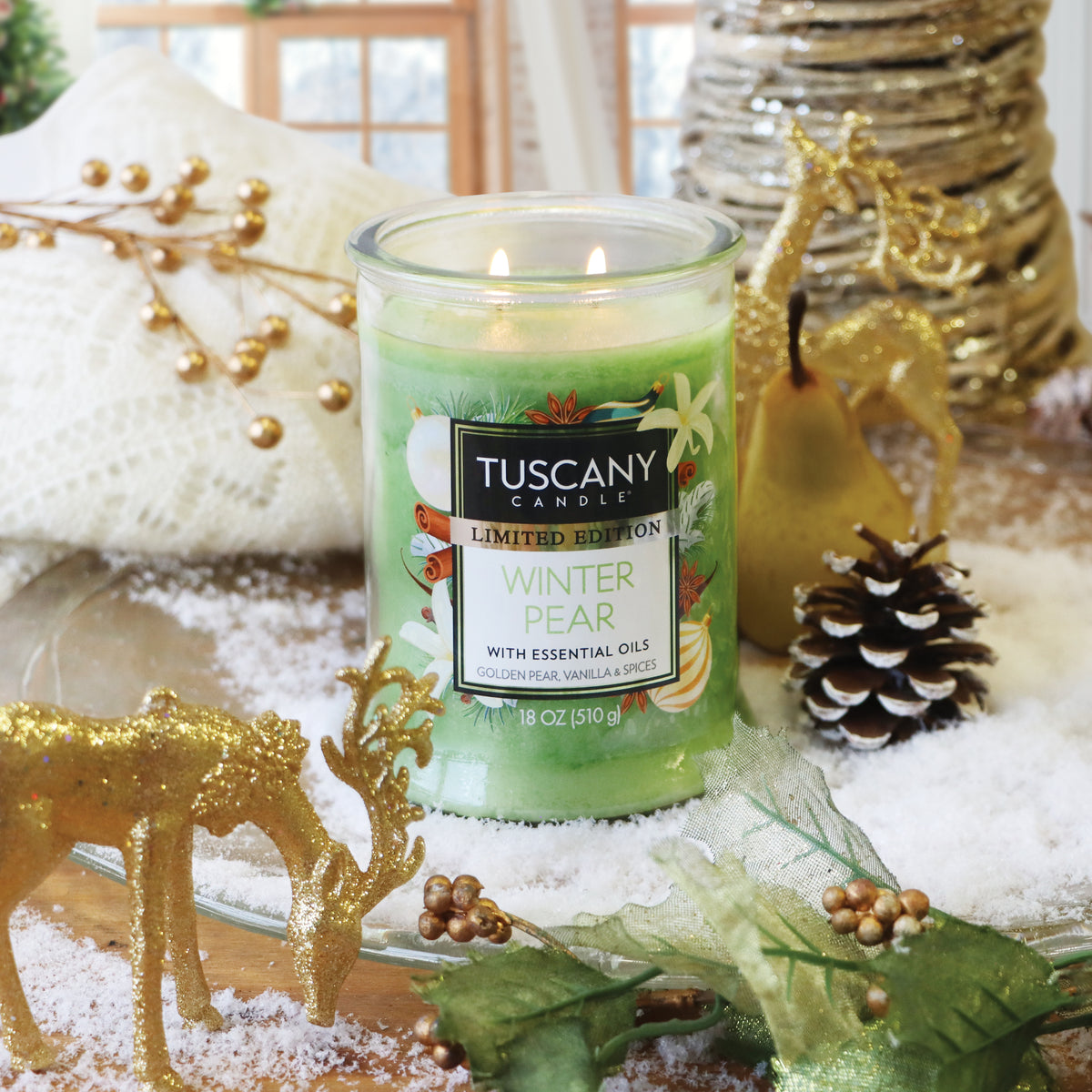 Tuscany Candle® SEASONAL Winter Pear: Golden Pear & Vanilla Scented Holiday Candle (18 oz) in a two-wick clear glass jar with green wax, surrounded by a pear, pinecone, and gold deer on artificial snow.