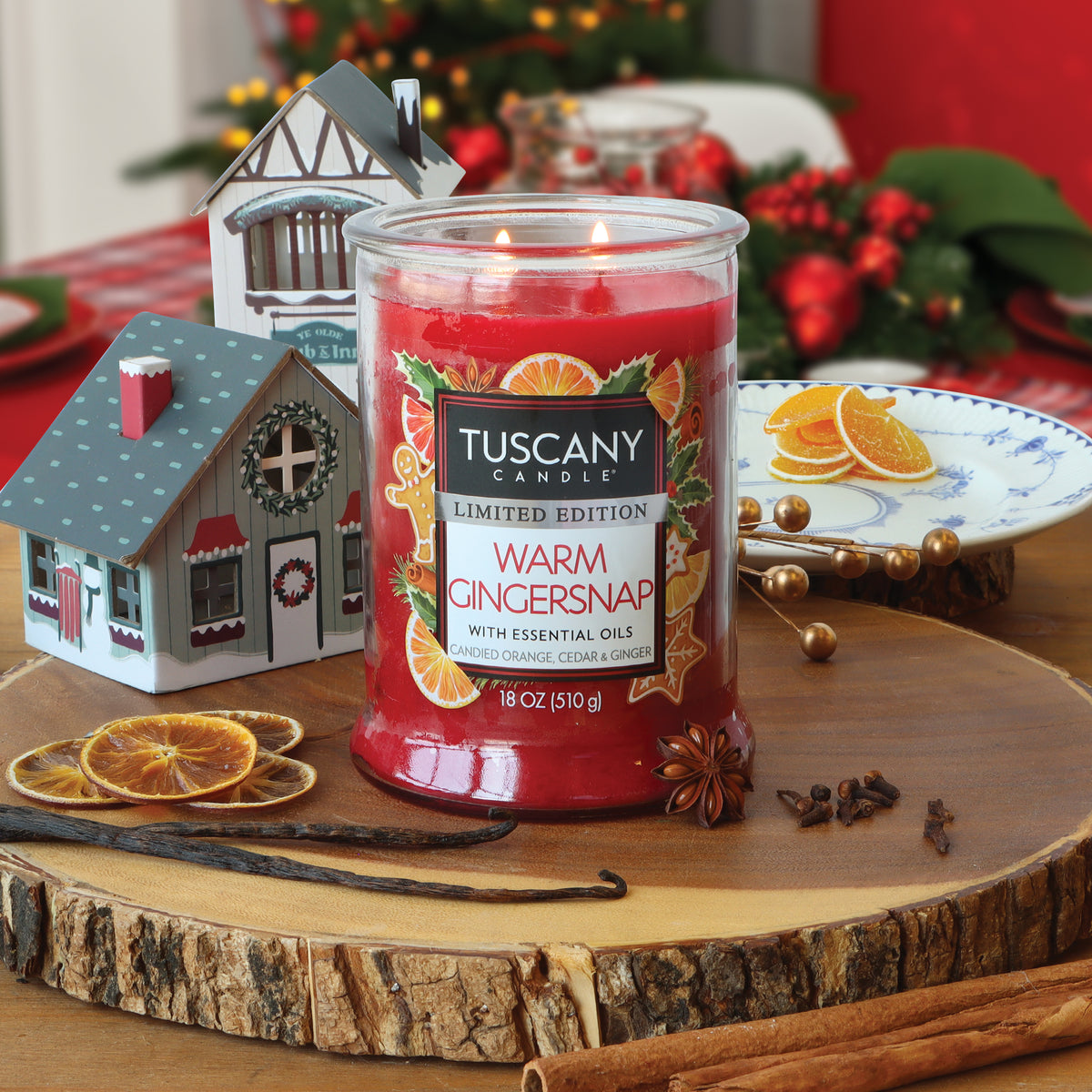 A red two-wick Warm Gingersnap: Candied Orange & Cedar Scented Holiday Candle (18 oz) by Tuscany Candle® sits on a wood slice decorated with dried orange slices, star anise, cloves, and vanilla beans.