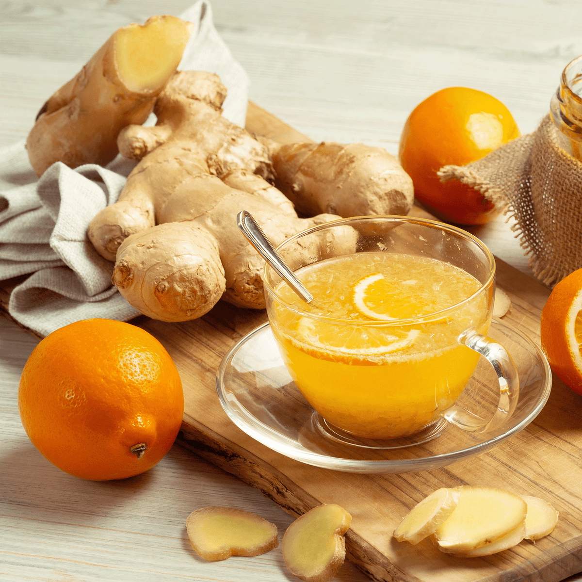 A glass teacup of citrus tea with a spoon, surrounded by ginger roots, sliced ginger, and fresh oranges on a wooden cutting board next to a Warm Gingersnap: Candied Orange & Cedar Scented Holiday Candle.