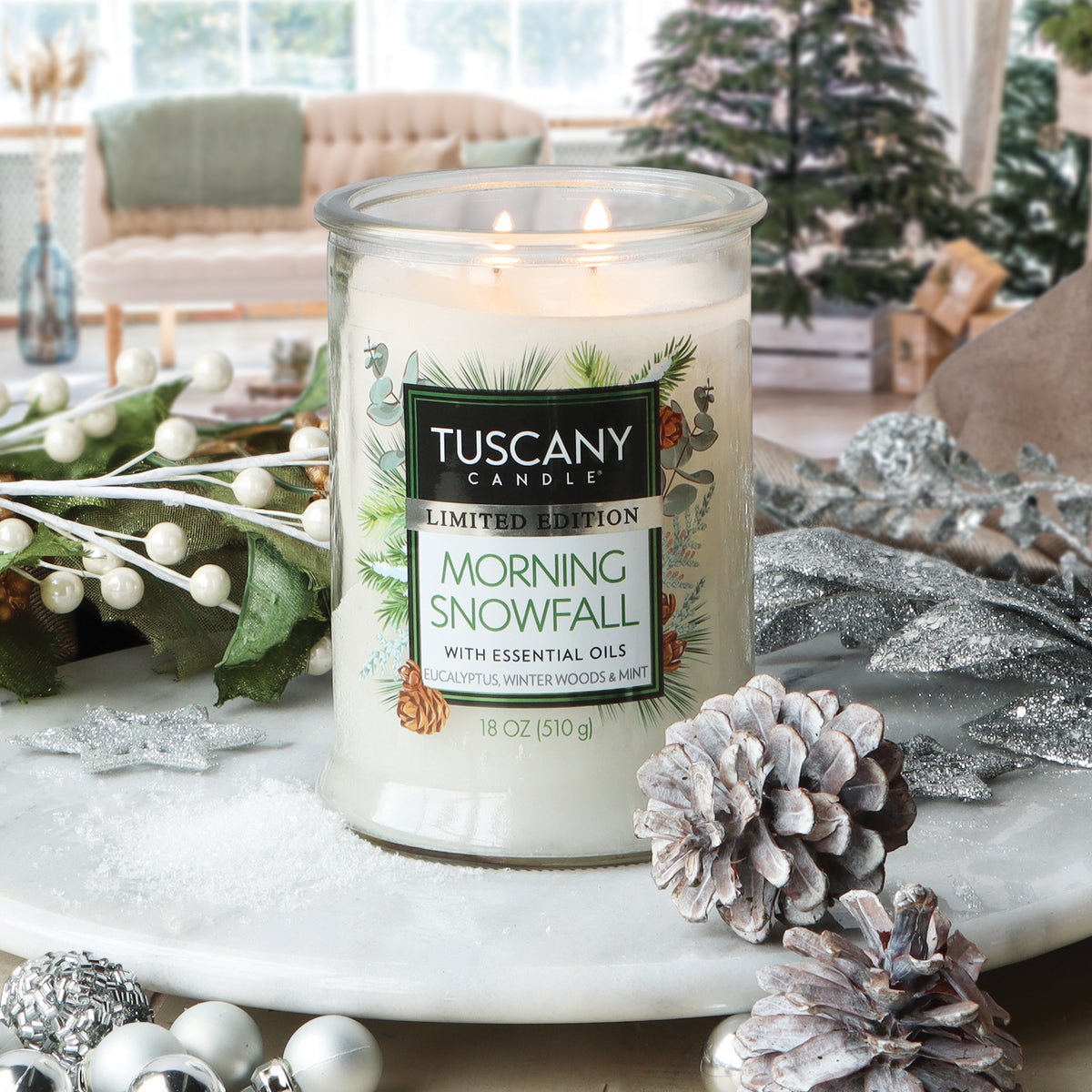A Tuscany Candle® SEASONAL Morning Snowfall: Eucalyptus & Winter Woods Scented Holiday Candle (18 oz) in a clear glass jar surrounded by pinecones, silver ornaments, and faux greenery on a white surface.