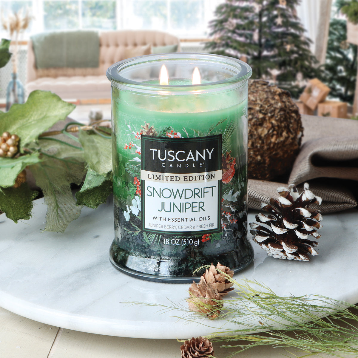 A lit green Tuscany Candle® SEASONAL Snowdrift Juniper: Juniper Berry & Fresh Pine Scented Holiday Candle (18 oz) in a glass jar, sitting on a marble surface with pinecones and greenery.