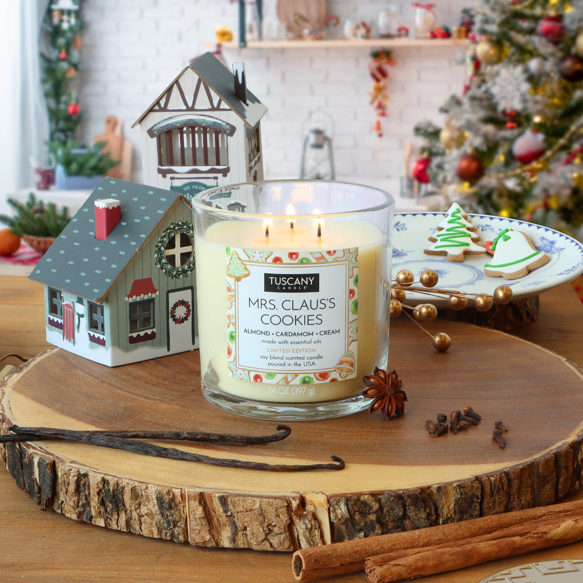 Mrs. Claus’s Cookies: Almond & Cardamom Scented Holiday Candle (14 oz) in a glass jar, surrounded by miniature houses, star anise, vanilla beans, and holiday cookies on a wooden board.