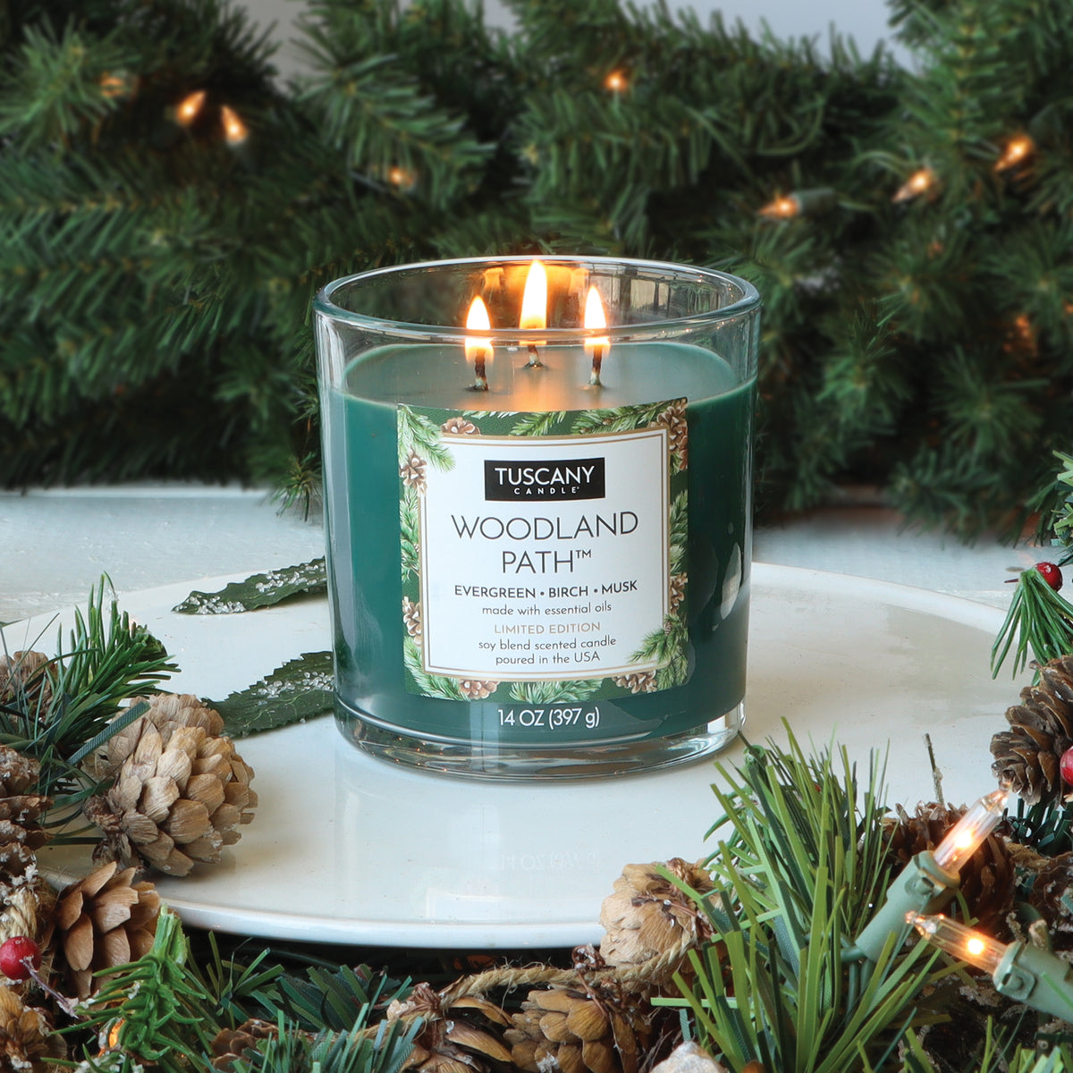 A lit, three-wick Tuscany Candle® SEASONAL Woodland Path®: Evergreen & Birch Scented Holiday Candle (14 oz) in a green glass jar, surrounded by pine branches, pinecones, and string lights.