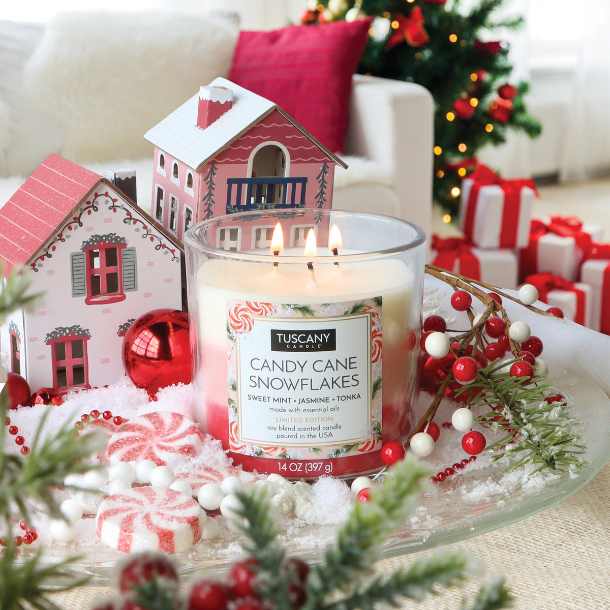 A 14 oz Tuscany Candle® SEASONAL Candy Cane Snowflakes: Sweet Mint & Jasmine Scented Holiday Candle in a glass jar, surrounded by miniature houses, peppermint candies, and snowy artificial pine branches.