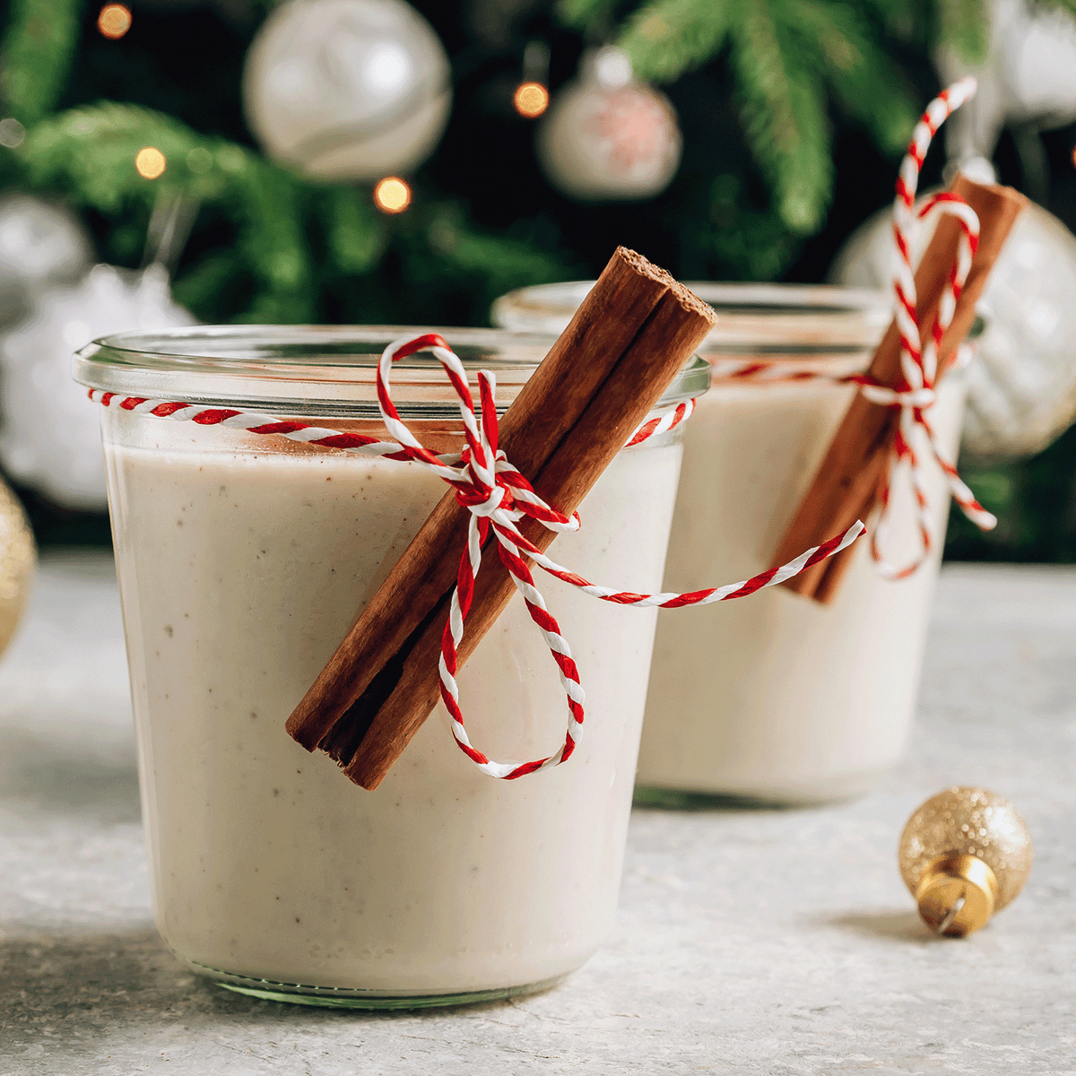 Two Santa’s Chai: Cinnamon & Clove Scented Holiday Candle (14 oz) glass jars filled with creamy beige wax, each tied with red and white twine and a cinnamon stick, against a blurred Christmas tree.
