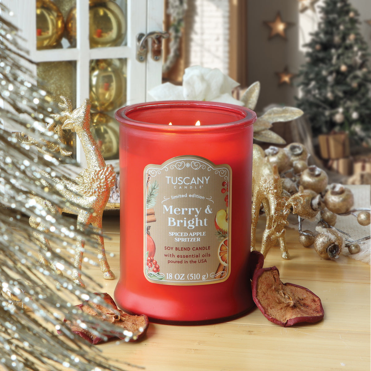 A lit red Tuscany Candle® SEASONAL Merry & Bright: Spiced Apple Spritzer Scented Holiday Candle (18 oz) surrounded by golden deer figures and dried apple slices.
