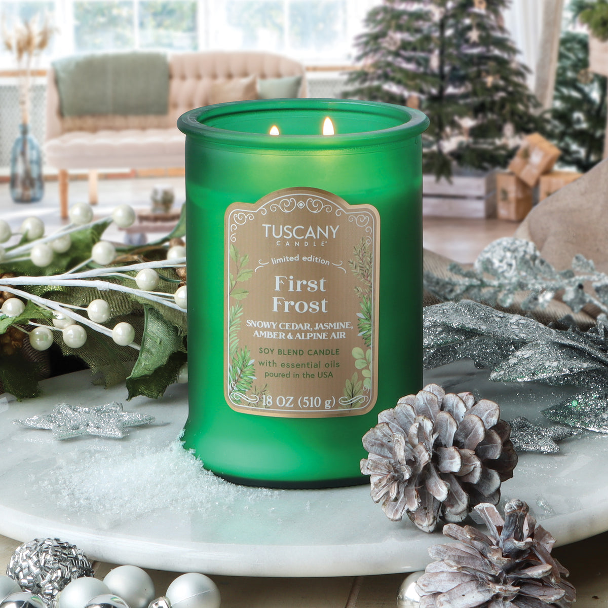 Green frosted glass First Frost: Jasmine & Cedar Scented Holiday Candle (18 oz) with two lit wicks on a white tray with pinecones and faux berries.