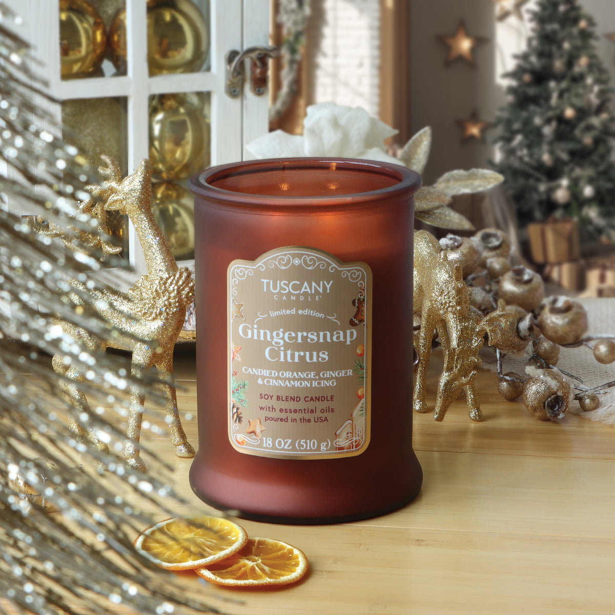 A brown glass jar of Tuscany Candle® SEASONAL Gingersnap Citrus: Candied Orange & Cinnamon Scented Holiday Candle (18 oz) sits on a table surrounded by gold holiday decorations and dried orange slices.