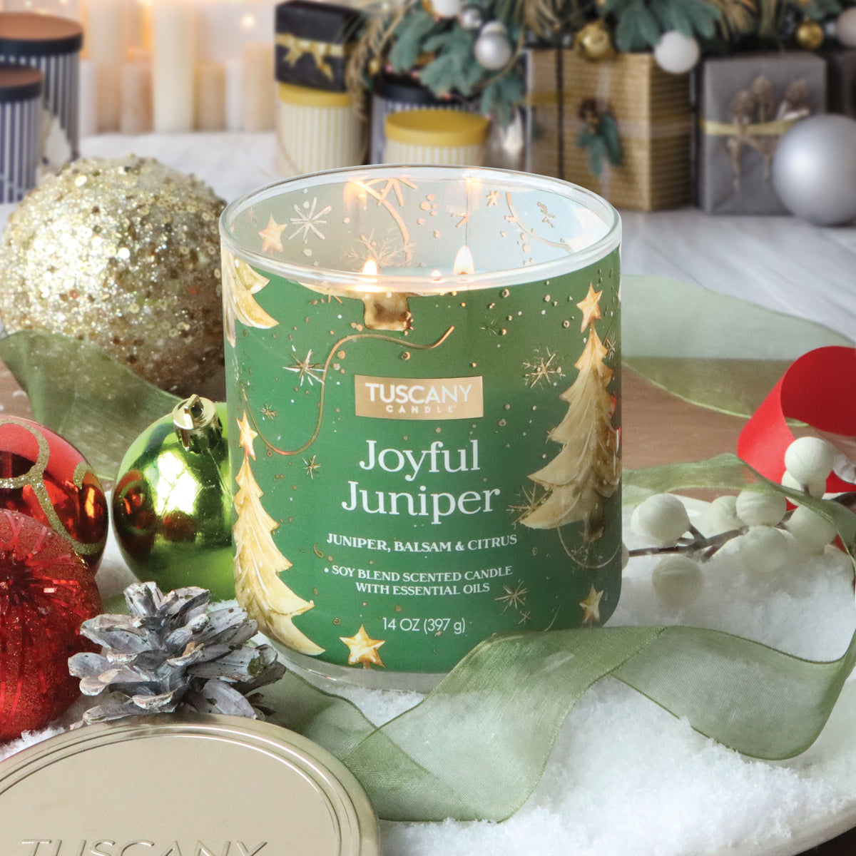 A Tuscany Candle® SEASONAL Joyful Juniper: Balsam & Citrus Scented Holiday Candle in a green glass jar with gold Christmas tree designs, surrounded by ornaments, a pinecone, and ribbon on artificial snow.