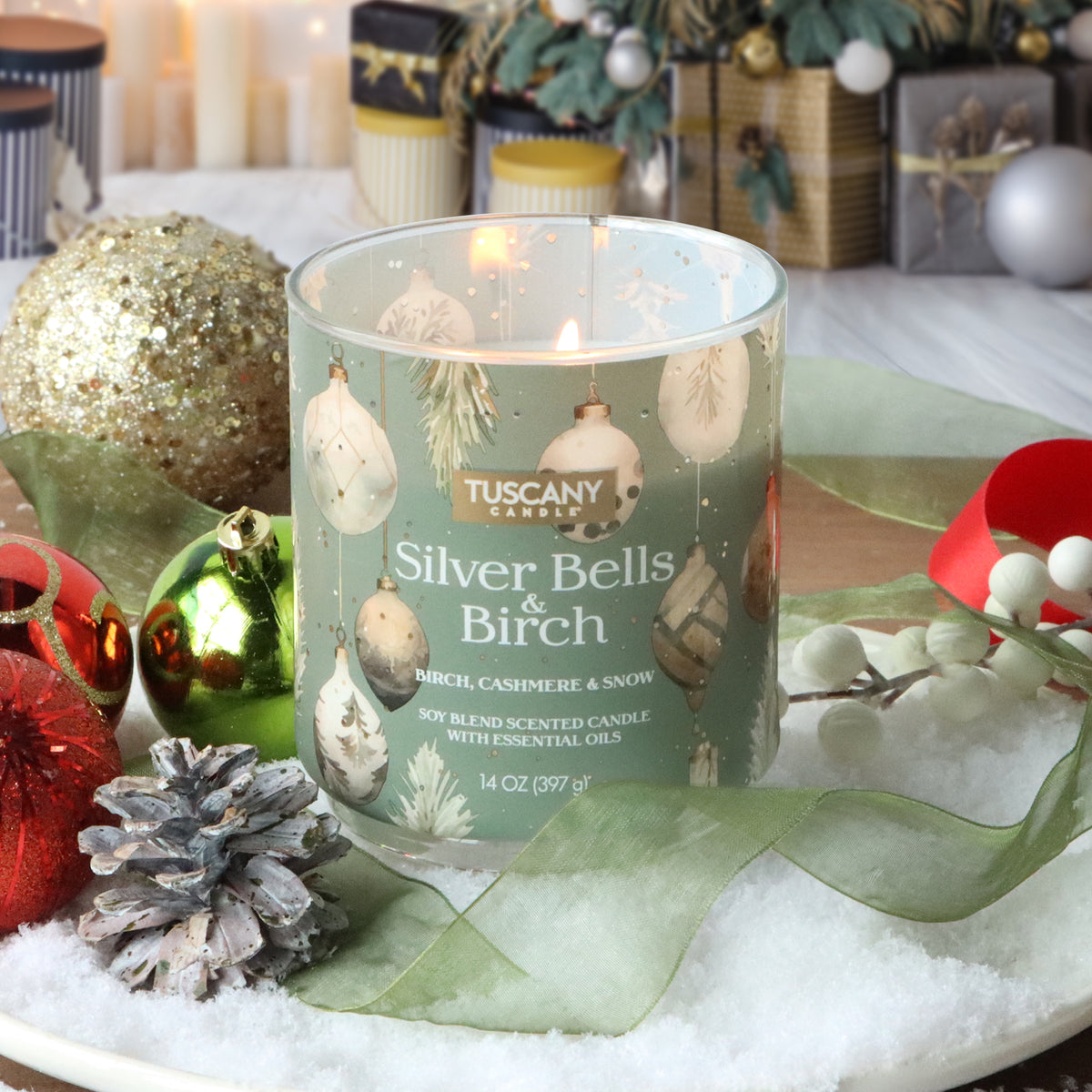 A Silver Bells & Birch: Cashmere & Snow Scented Holiday Candle (14 oz) in a glass jar, decorated with holiday ornaments on a bed of artificial snow with pinecones and green ribbon.