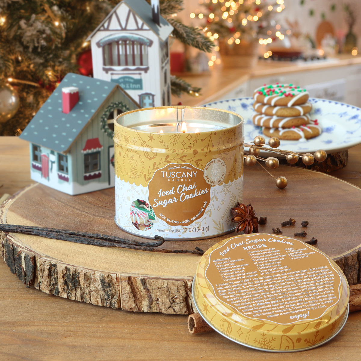 A 12 oz Tuscany Candle® SEASONAL Iced Chai Sugar Cookies: Vanilla & Buttercream Scented Holiday Candle sits on a wooden slice with a vanilla bean, star anise, a cinnamon stick, and a recipe lid.
