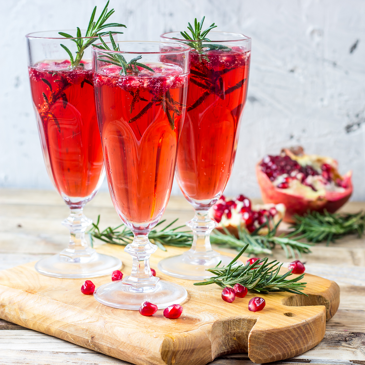 Three glasses of Merry Mimosa: Pomegranate & Rhubarb Scented Holiday Candle (12 oz) red pomegranate cocktail garnished with fresh rosemary sprigs on a wooden board with pomegranate seeds.