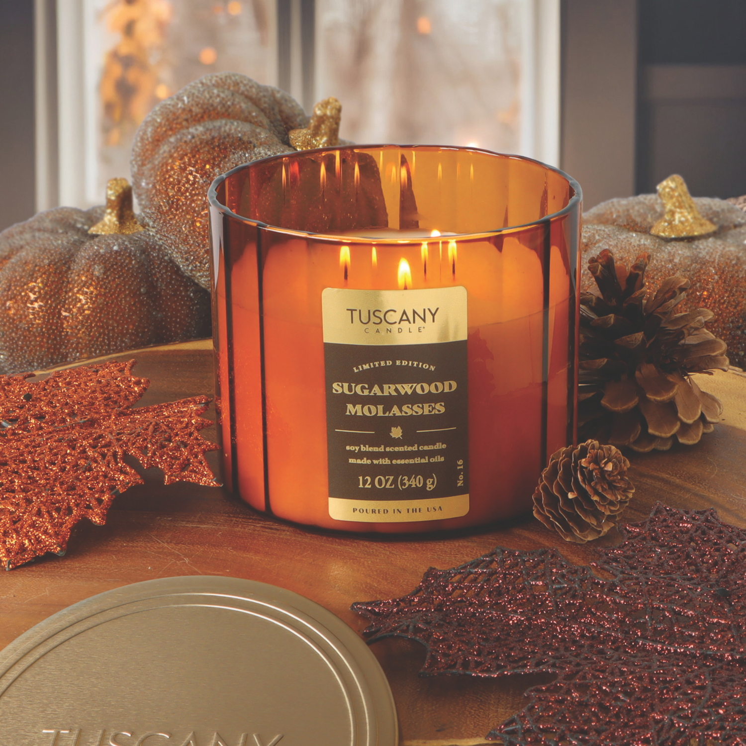 A Tuscany Candle® SEASONAL Sugarwood Molasses: Maple & Chestnut Scented Fall Candle (12 oz) sits on a table with pinecones, pumpkins, and autumn leaves in a cozy autumn arrangement.