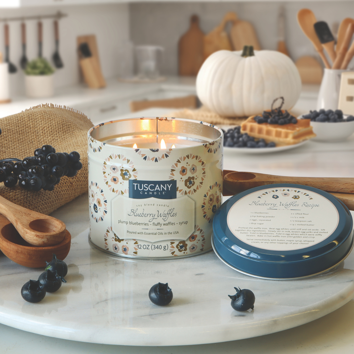 A lit Tuscany Candle® SEASONAL Blueberry Waffles: Maple Syrup & Waffle Scented Fall Candle (12 oz, Bakery Tins Collection) sits on a marble tray with fresh blueberries, a candle lid, wooden utensils, and a white pumpkin in the kitchen.