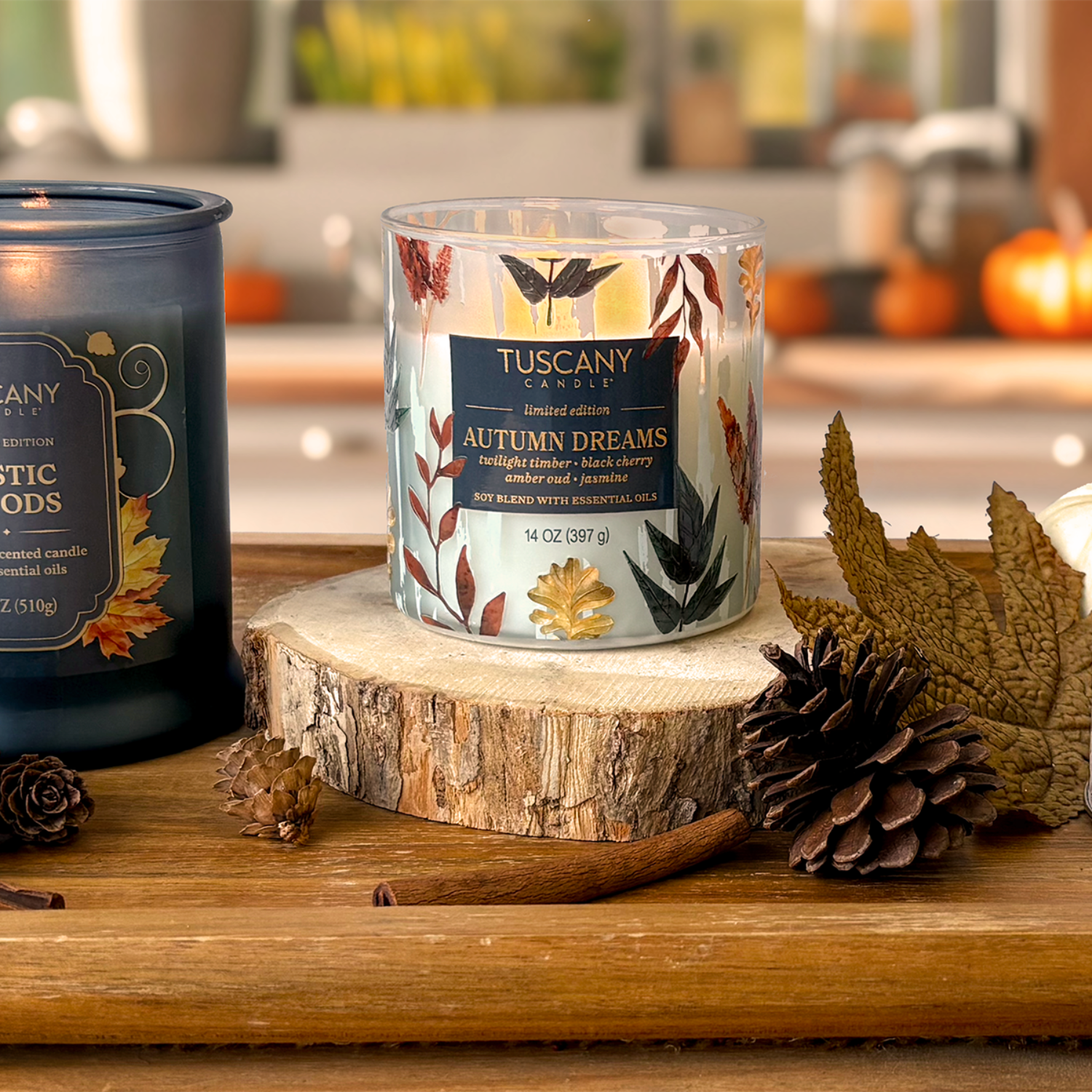 The Tuscany Candle® SEASONAL Autumn Dreams: Black Cherry & Amber Oud Scented Fall Candle (14 oz) from the Simply Fall Collection sits on a wooden slice with pinecones and leaves; another candle is nearby, with a blurred kitchen in the background.