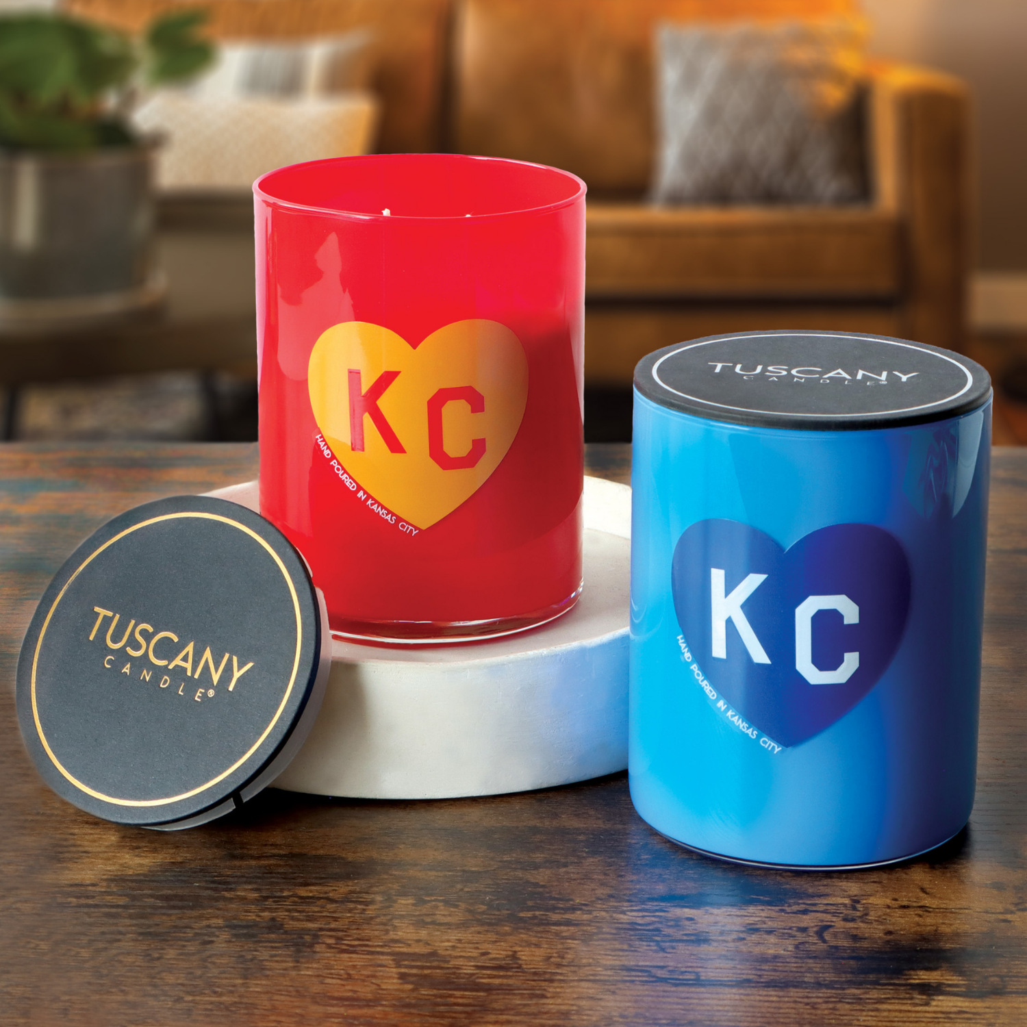Two 13oz Fall Festival Scented Red KC Heart Candles by Tuscany Candle x Charlie Hustle are displayed on a wooden table, highlighting their signature "KC" heart design and unique collaboration.