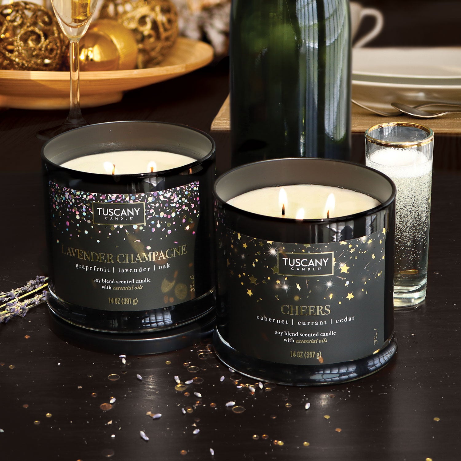 A Cheers Scented Candle (14 oz) from the Tuscany Candle brand on a table next to a bottle of champagne for a holiday celebration.