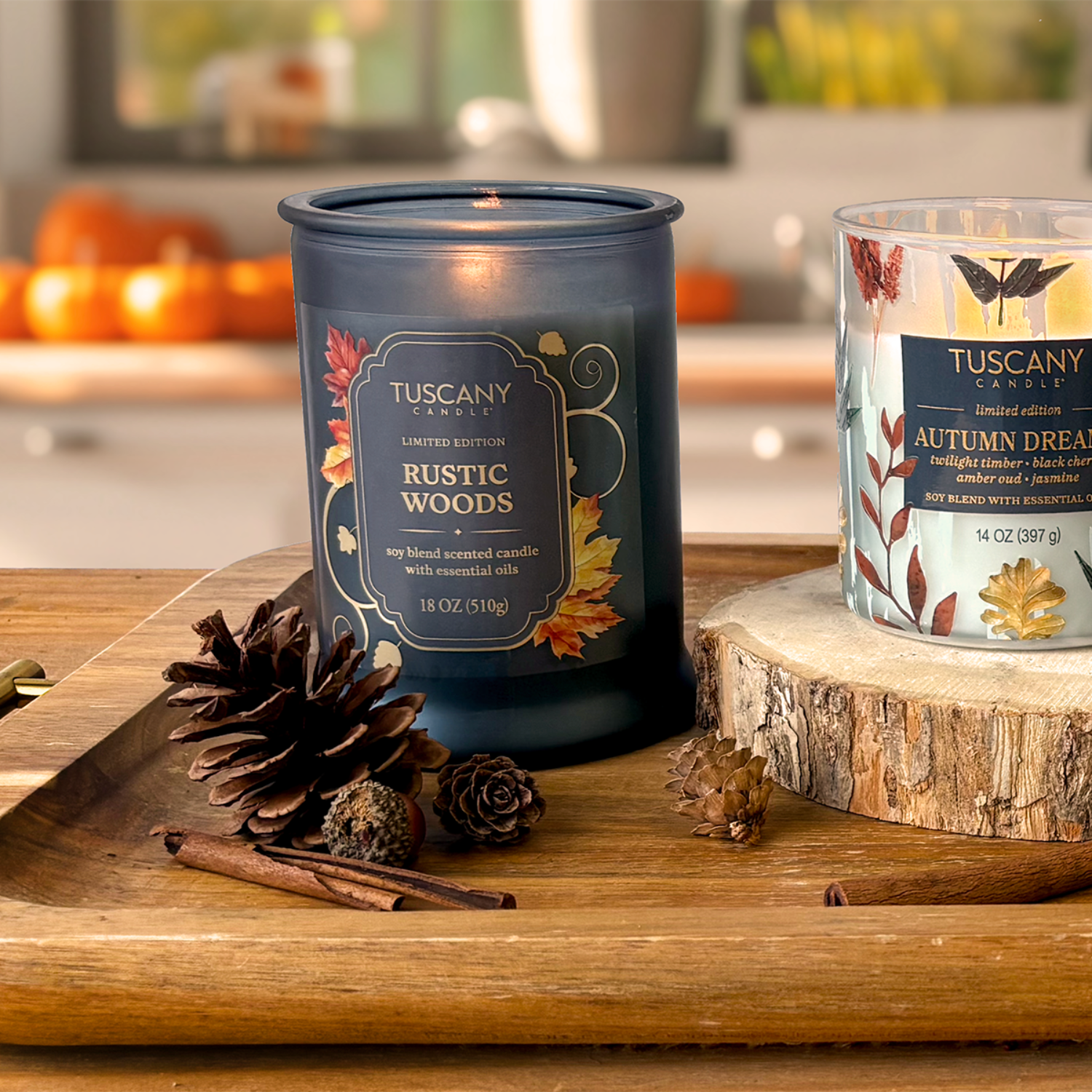 A blue Tuscany Candle® SEASONAL Rustic Woods: Sandalwood & Vanilla Scented Fall Candle (18 oz) and a clear Autumn Dreams soy wax blend candle sit on a wooden tray with pinecones and a wood slice in an autumn-themed kitchen.