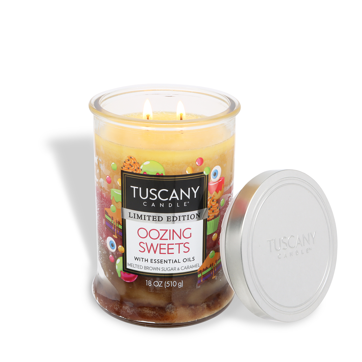 An 18 oz glass jar candle labeled "Oozing Sweets: Brown Maple Sugar & Caramel" by Tuscany Candle® SEASONAL features two lit wicks. The "Tuscany Candle" lid rests beside the jar, enhancing the Halloween ambiance with a sweet caramel scent.