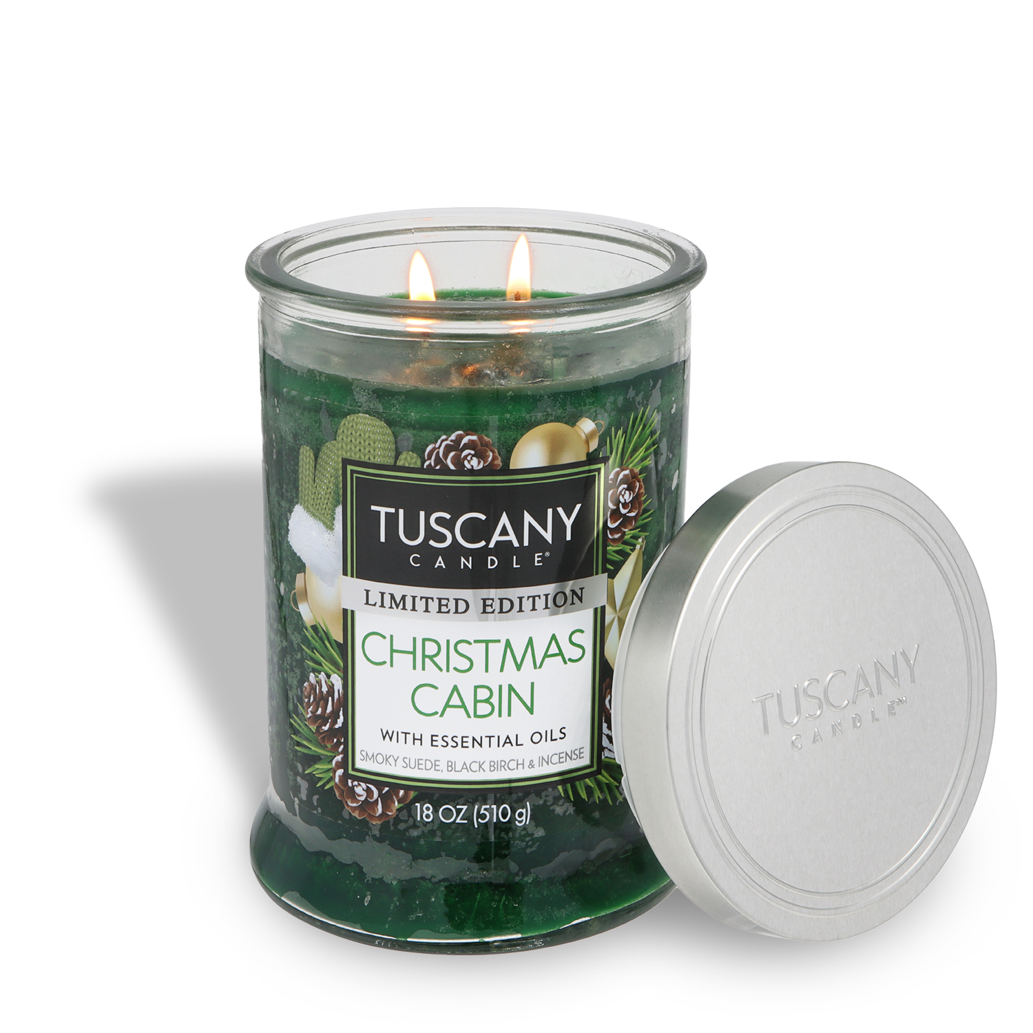 An 18 oz green glass jar candle labeled "Christmas Cabin: Smoky Suede & Birch" by Tuscany Candle® SEASONAL, with two wicks burning and a silver lid beside it.