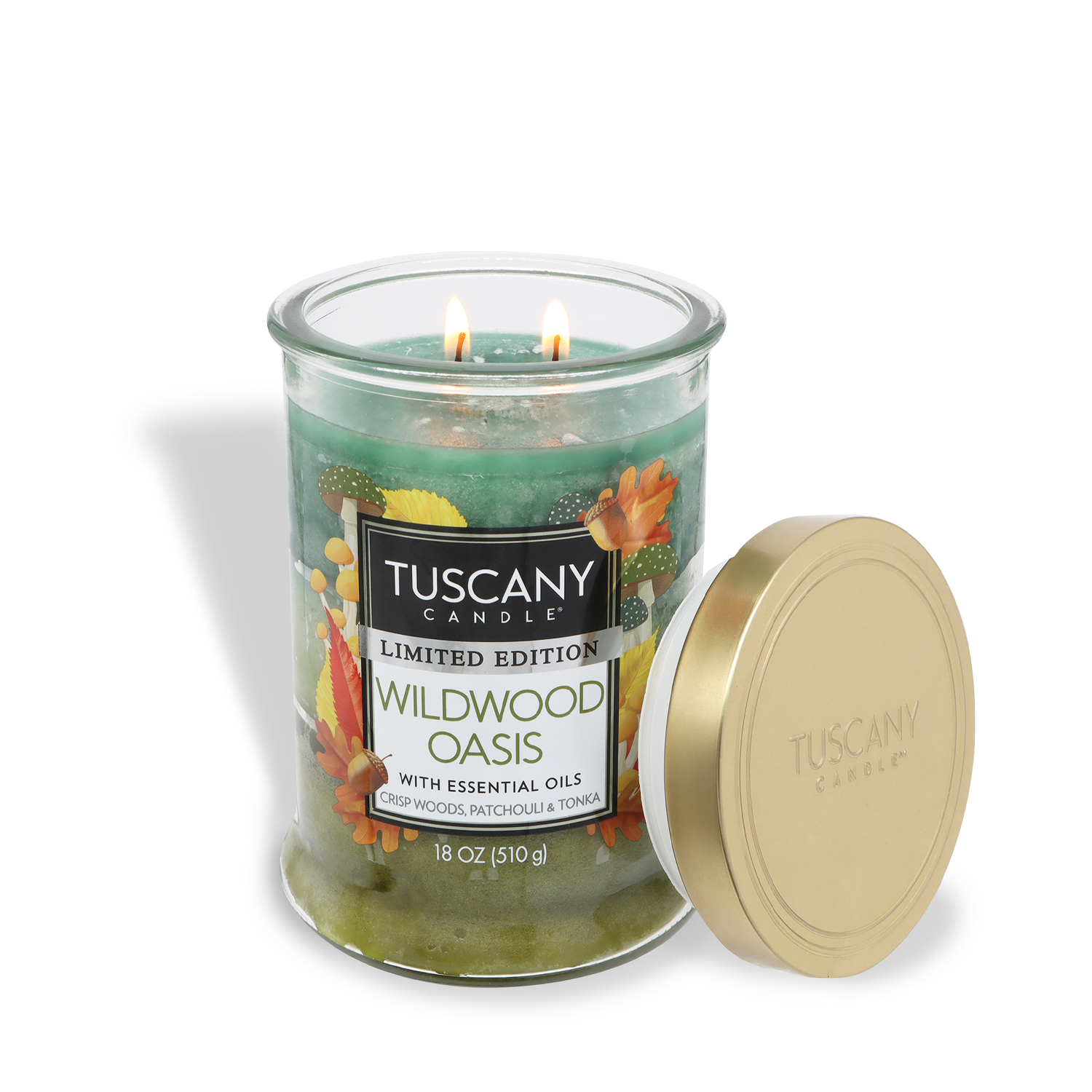 An 18 oz Tuscany Candle® SEASONAL "Wildwood Oasis: Amber Woods & Patchouli" jar candle with two lit wicks and a gold lid beside it. The label displays colorful autumn leaves and greenery.