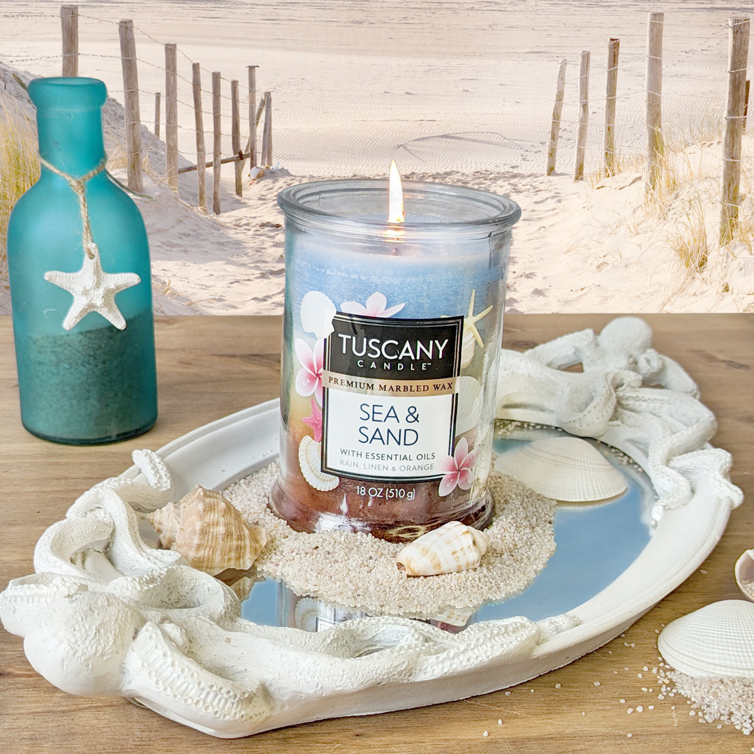 A Sea & Sand Long-Lasting Scented Jar Candle (18 oz) by Tuscany Candle® EVD nestled on a tray with shells and sand.