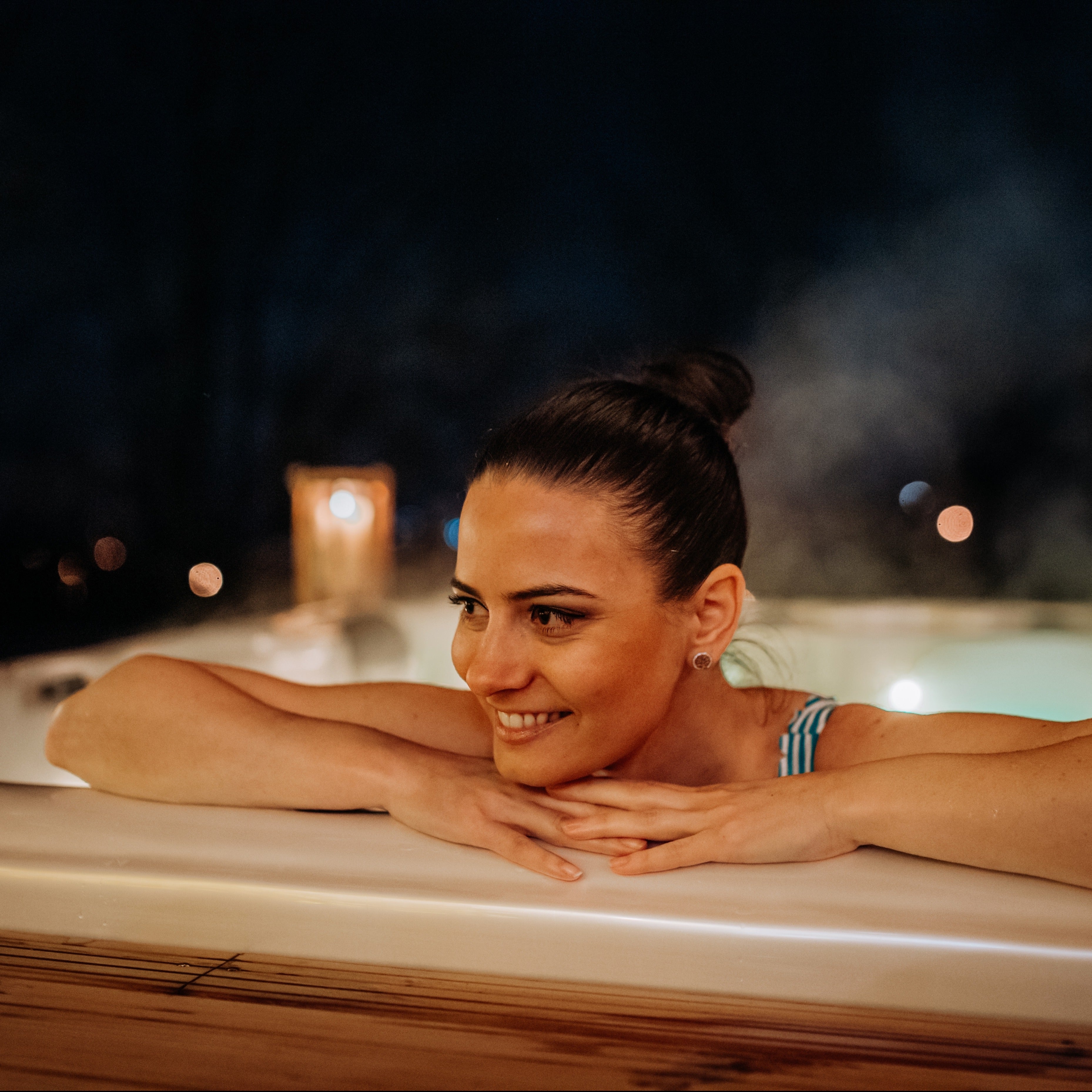 A woman relaxing in a Tuscany Candle-lit hot tub at night, promoting Winter Wellness and embracing the soothing ambiance of the Amber Meditation Scented Jar Candle (13oz) from the Winter Wellness Collection.